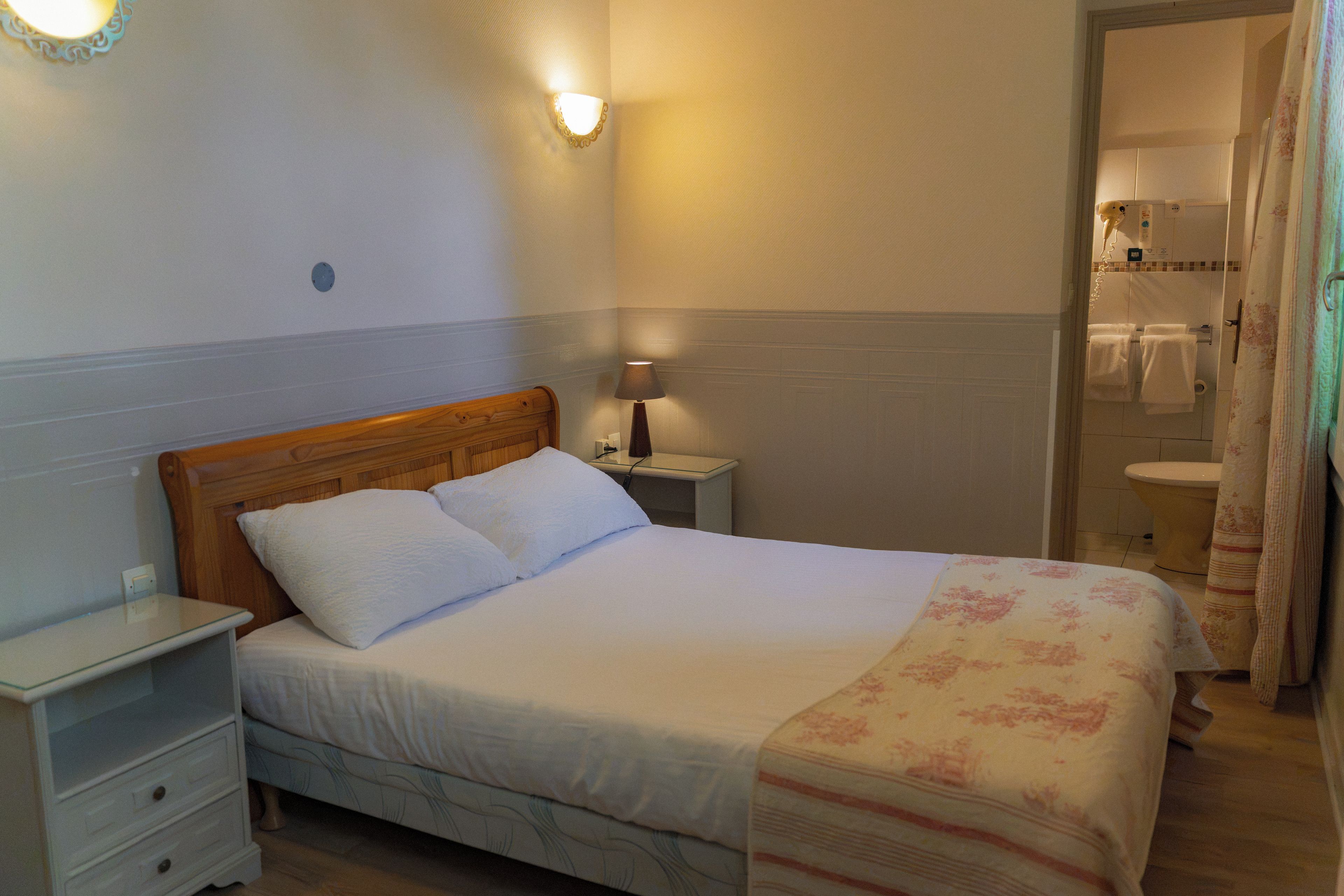 Une chambre d’hôtel avec un lit, une table de chevet, une lampe et une salle de bain visible à travers une porte ouverte.