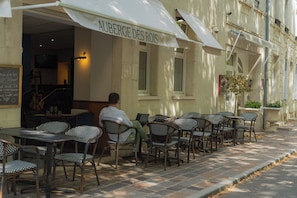 Un café en plein air avec des chaises et des tables en osier, un tableau noir servant de menu, et un bâtiment sur lequel on peut lire l’inscription « AUBERGE DES ROIS ».