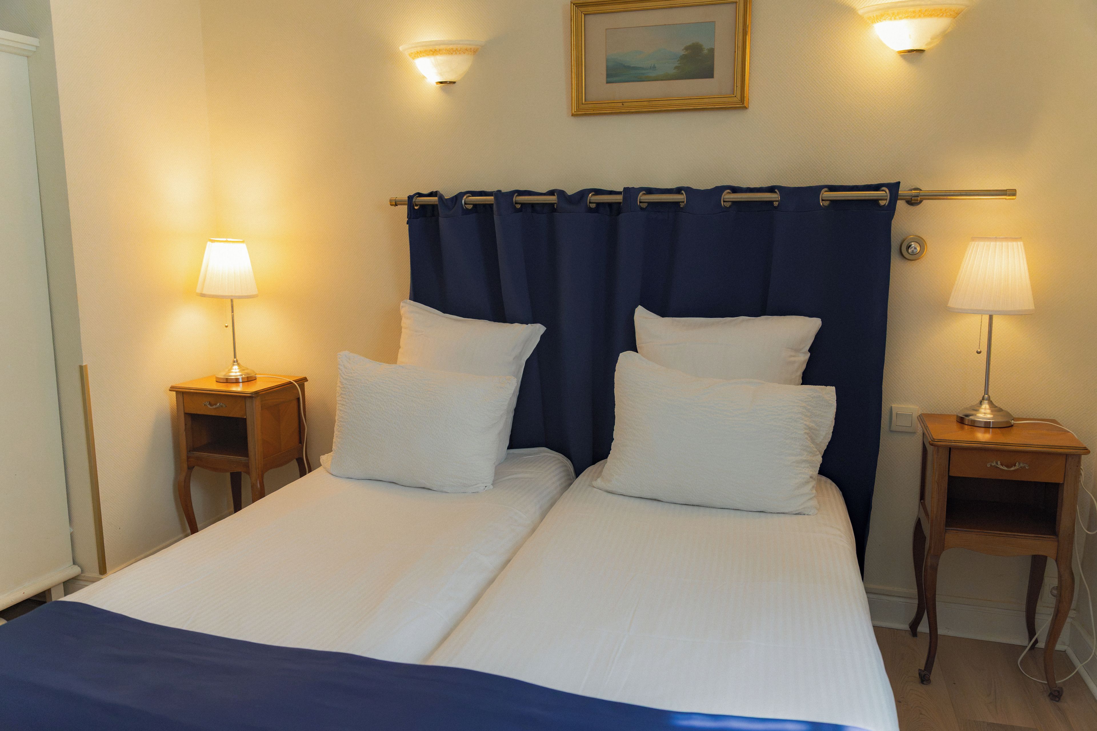 Uma cama de casal com duas almofadas brancas, uma cortina azul escura e duas luminárias de cabeceira.