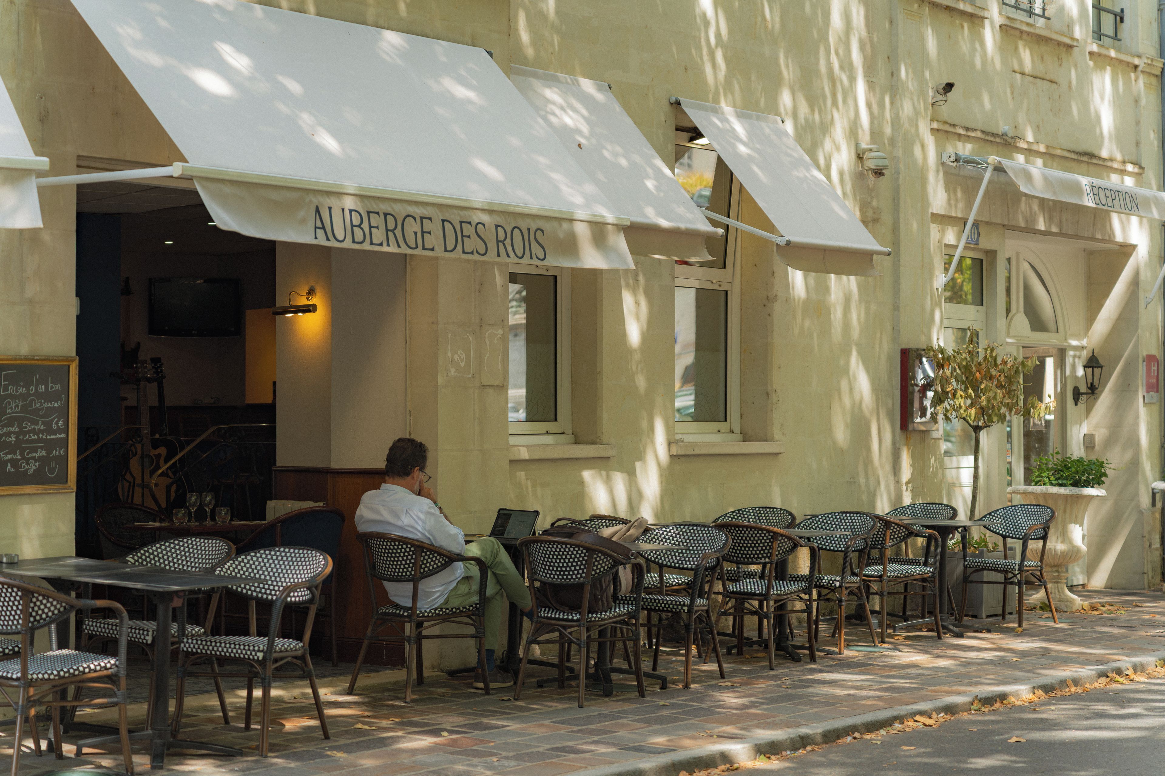 Un café al aire libre con un letrero que dice 'AUBERGE DES ROIS' y un hombre sentado en una mesa.