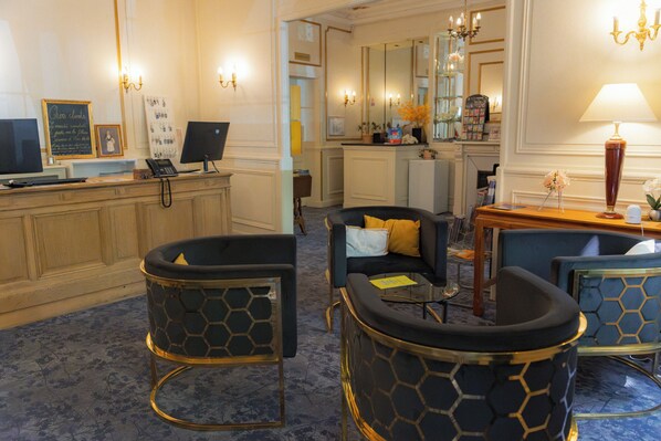 Un hall d’hôtel comprenant un comptoir d’accueil, un espace salon avec des fauteuils, une table basse et un lustre.