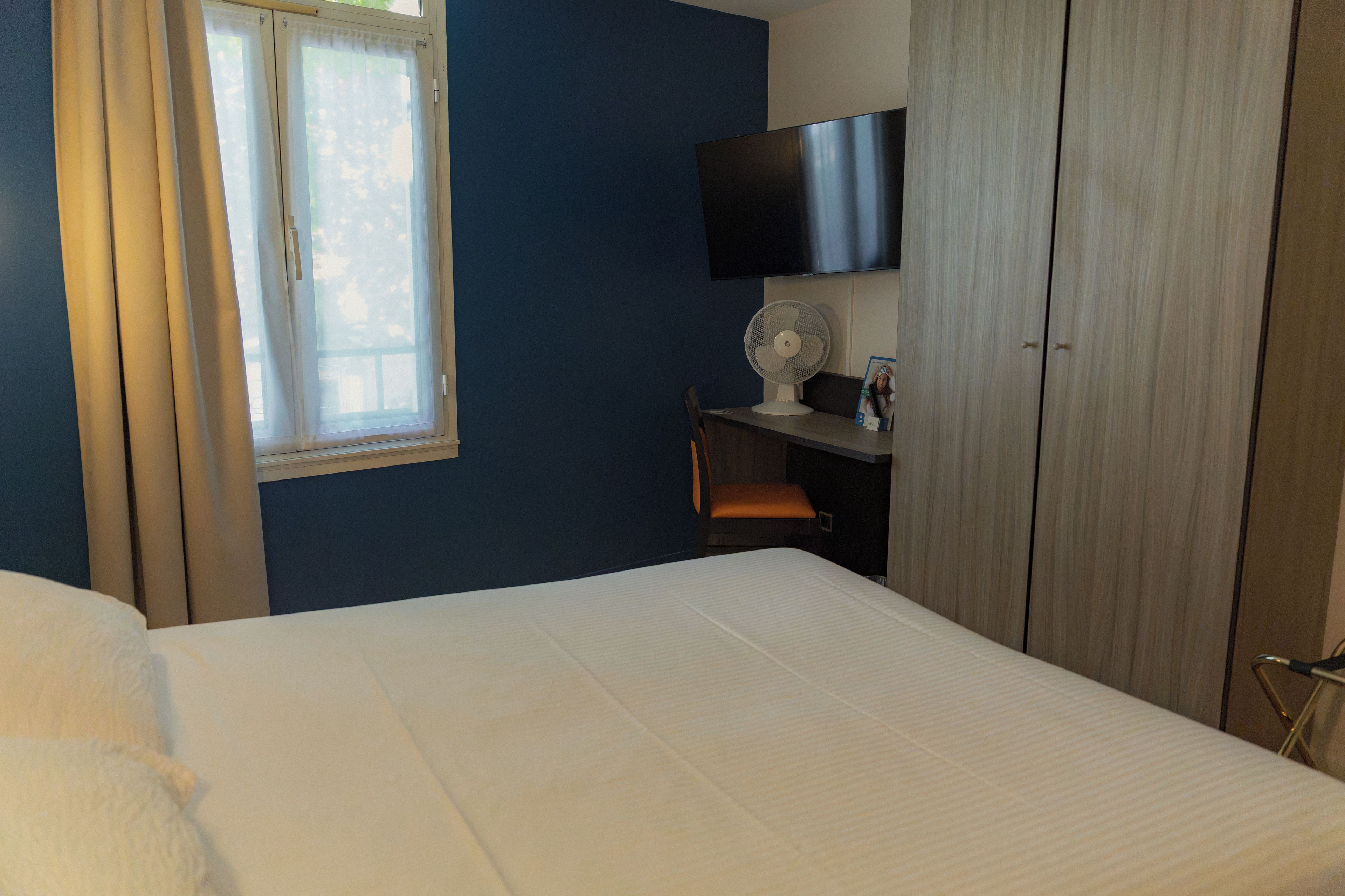A hotel room with a bed, a desk with a chair, a television, and a window with curtains.