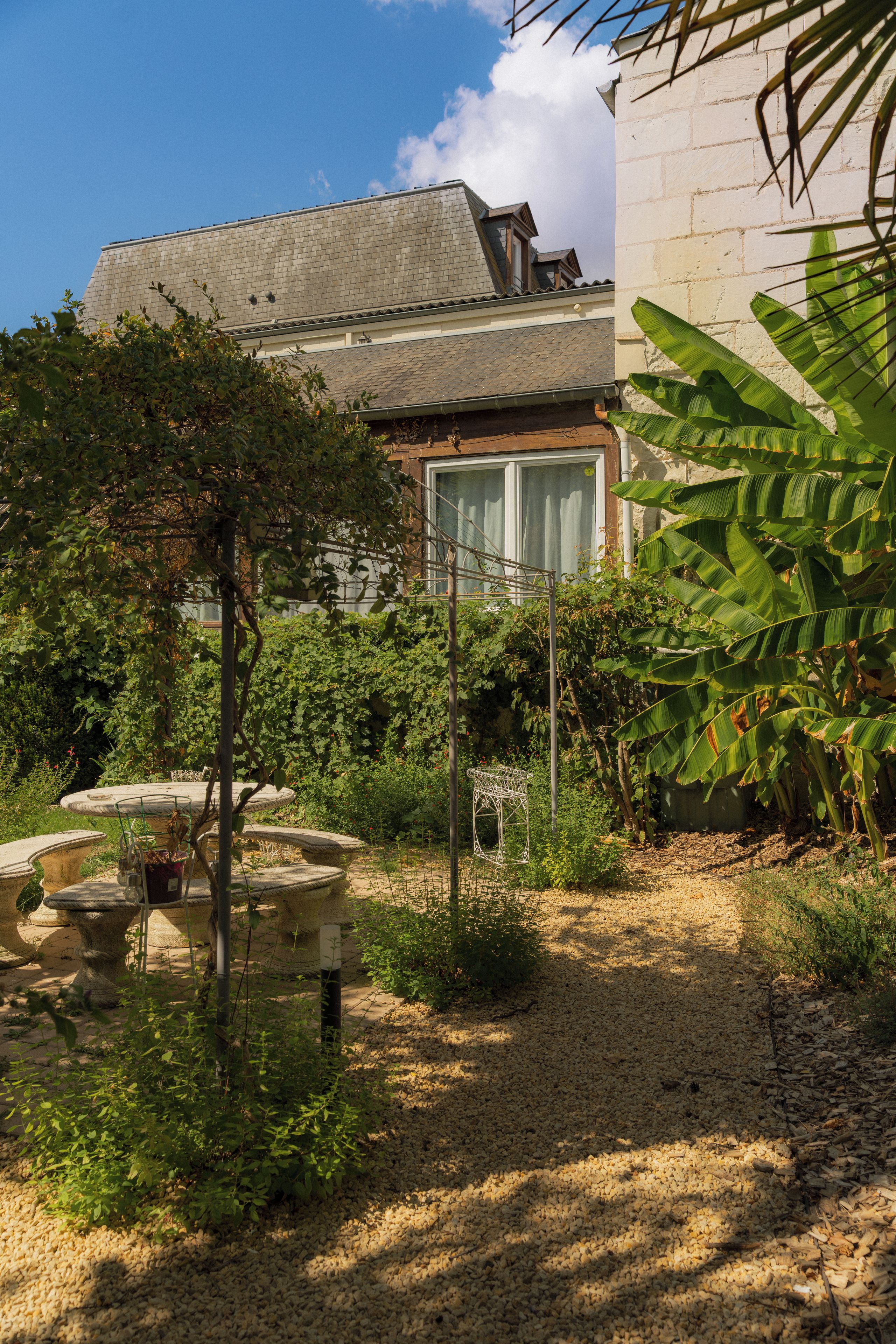 Un jardín con un banco de piedra, un árbol pequeño y una casa con una ventana al fondo.