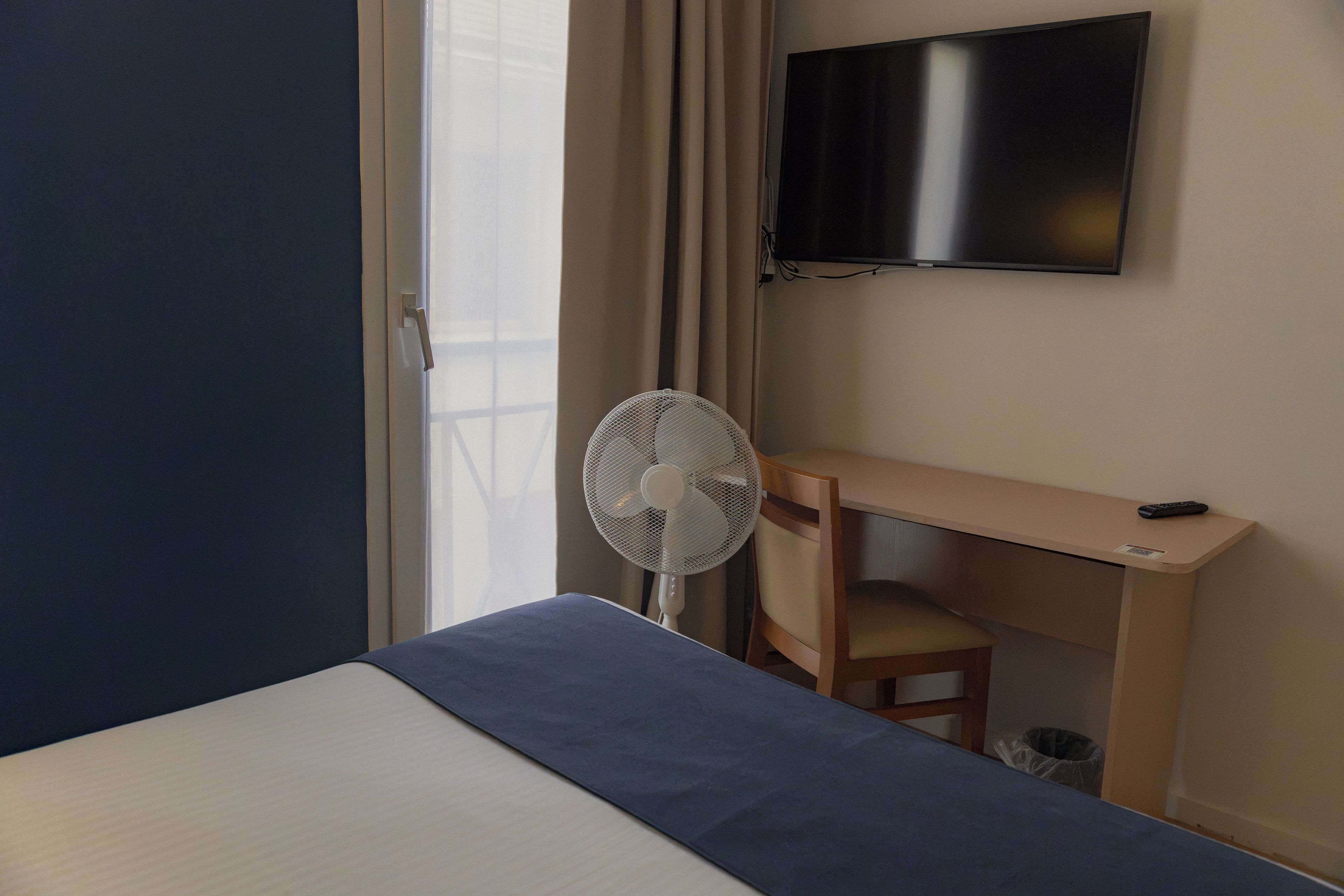 A hotel room with a bed, a desk, a chair, a TV, and a standing fan.