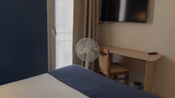 A hotel room with a bed, a desk, a chair, a TV, and a standing fan.