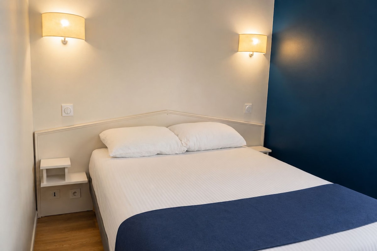 Een net opgemaakt bed met wit bedgoed en een blauw accent, met aan beide kanten nachtkastjes met lampen, in een kamer met een effen witte muur en een donkerblauwe accentmuur.