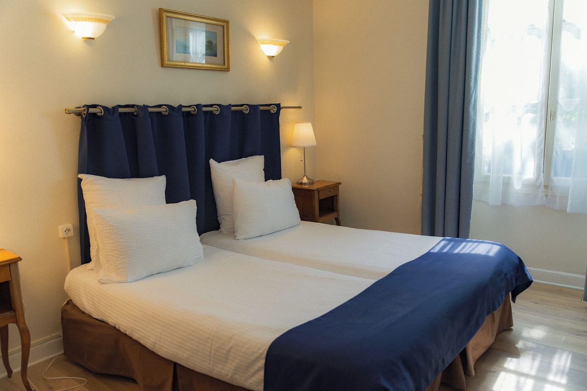 Een tweepersoonsbed met witte lakens en een blauwe sprei, een nachtkastje met een lamp, een aan de muur bevestigd fotolijstje, en een raam met gordijnen.