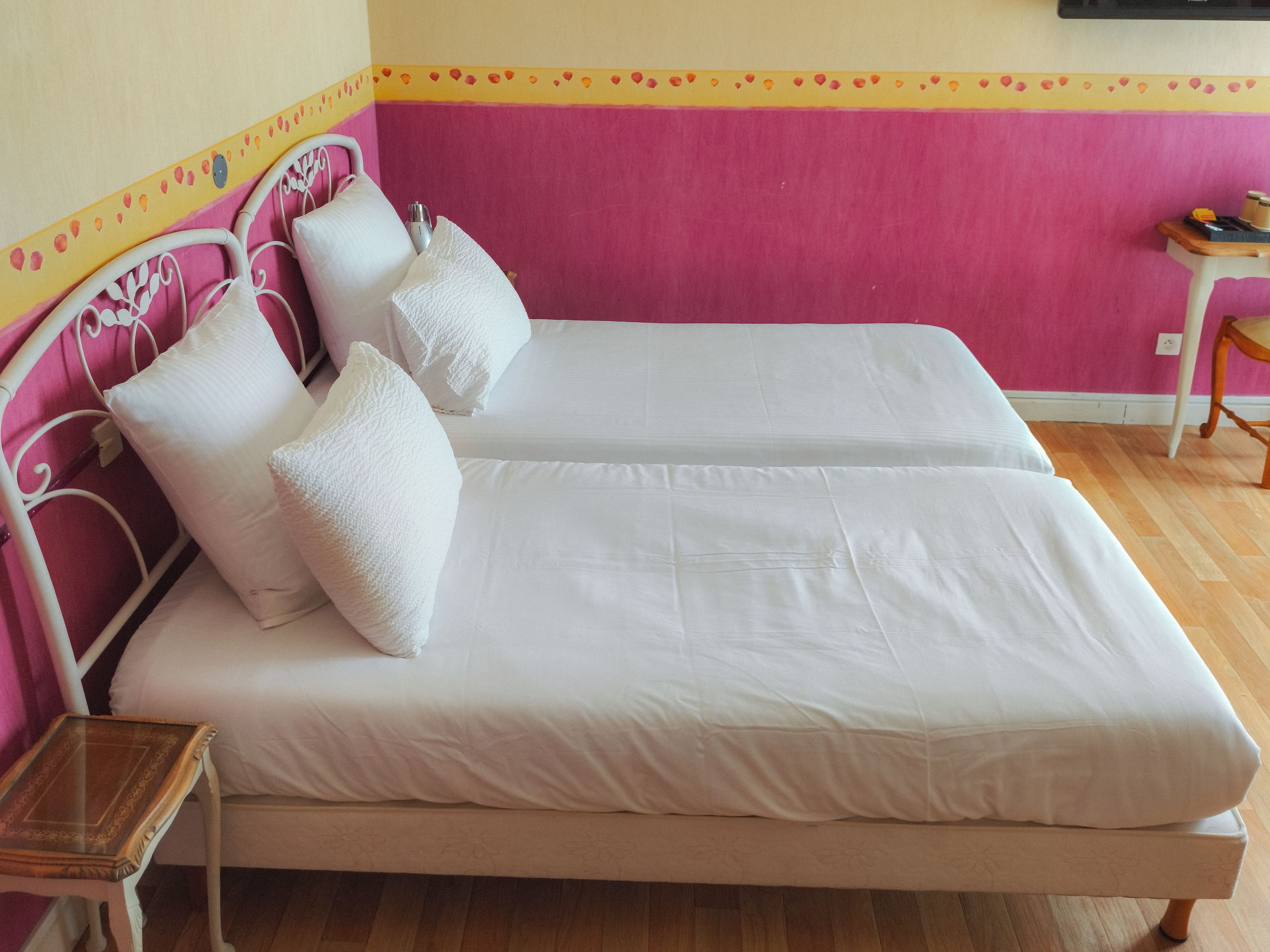 A double bed with white linens and pillows, a bedside table with a glass top, and a wooden chair.