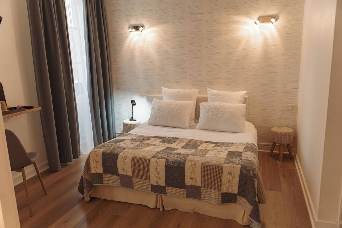 Une chambre à coucher avec un grand lit, une table de chevet, une lampe, une chaise et une fenêtre avec des rideaux.