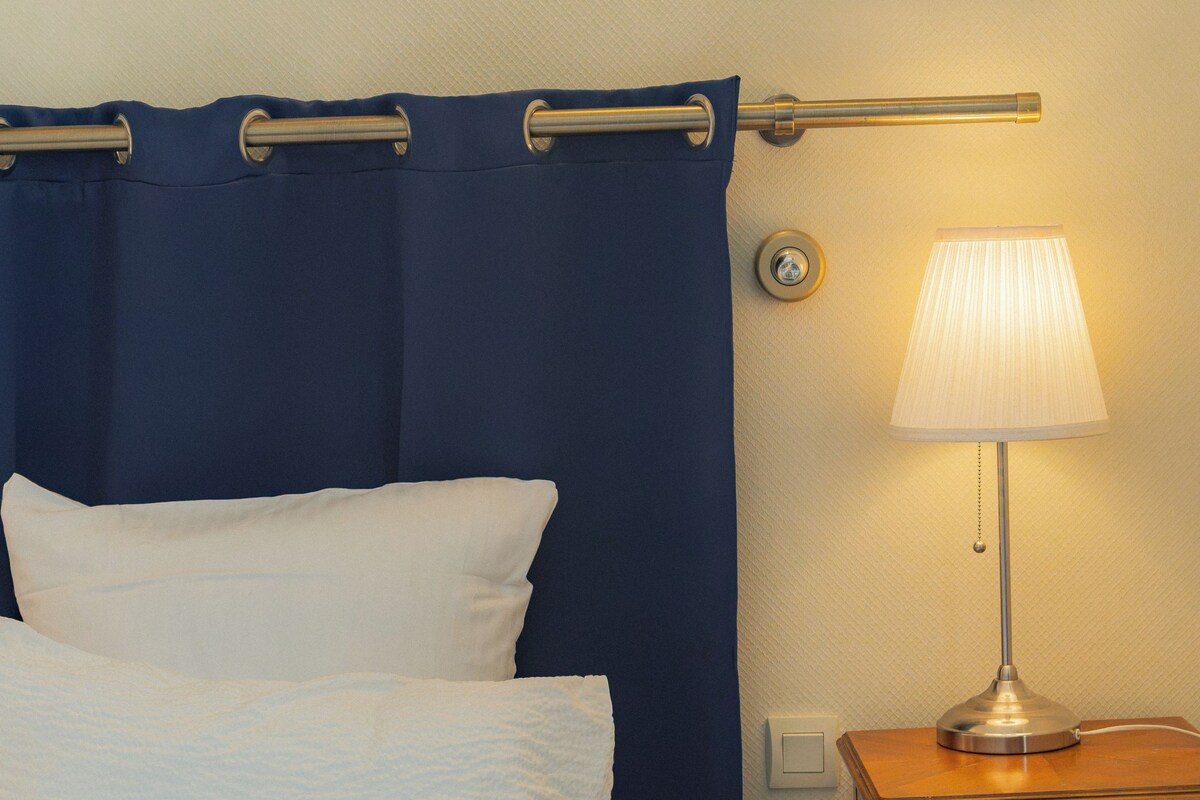 Un lit avec du linge de lit blanc, un rideau bleu, une lampe de chevet et une table de chevet en bois avec un tiroir.