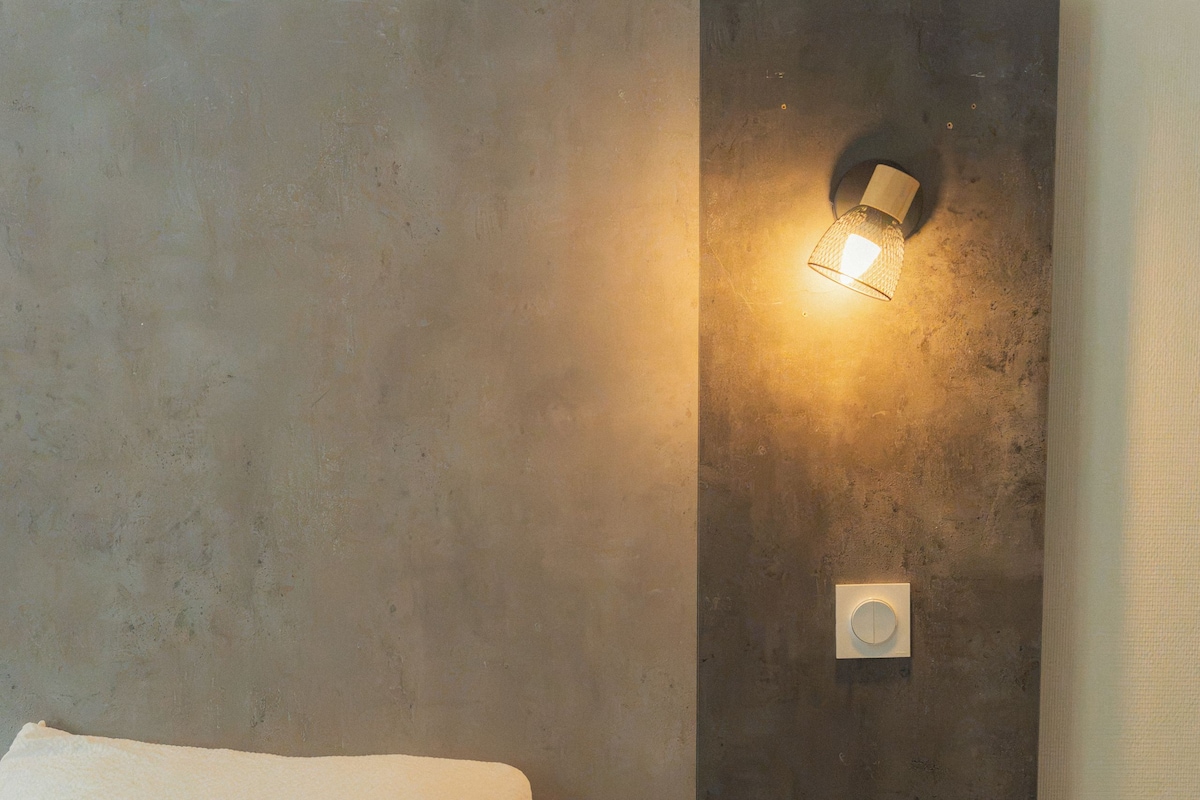 Een slaapkamer met een bed, een nachtkastje, een wandlamp en een stopcontact.