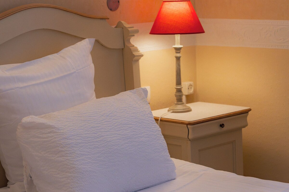 Un lit avec du linge de lit blanc et des oreillers, une table de chevet avec une lampe rouge et un luminaire fixé au mur.