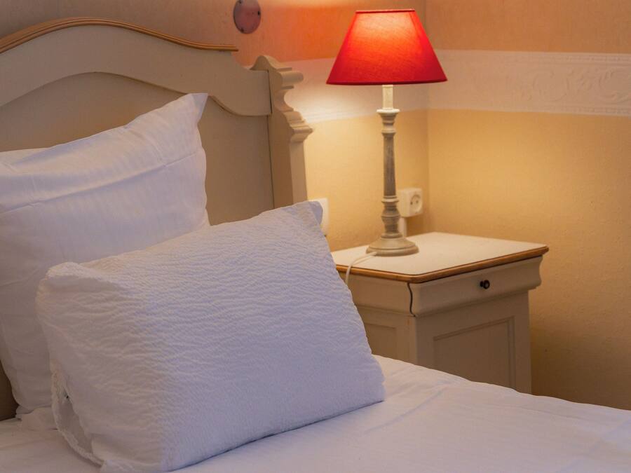 A bed with white linens and pillows, a bedside table with a red lamp, and a wall-mounted light fixture.