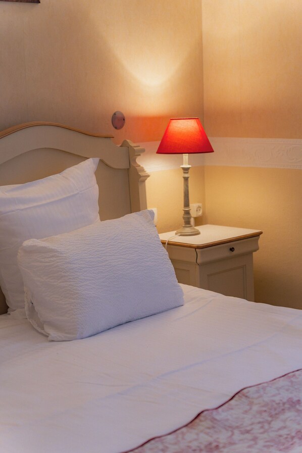 A bed with white linens and pillows, a bedside table with a red lamp, and a wall-mounted light fixture.