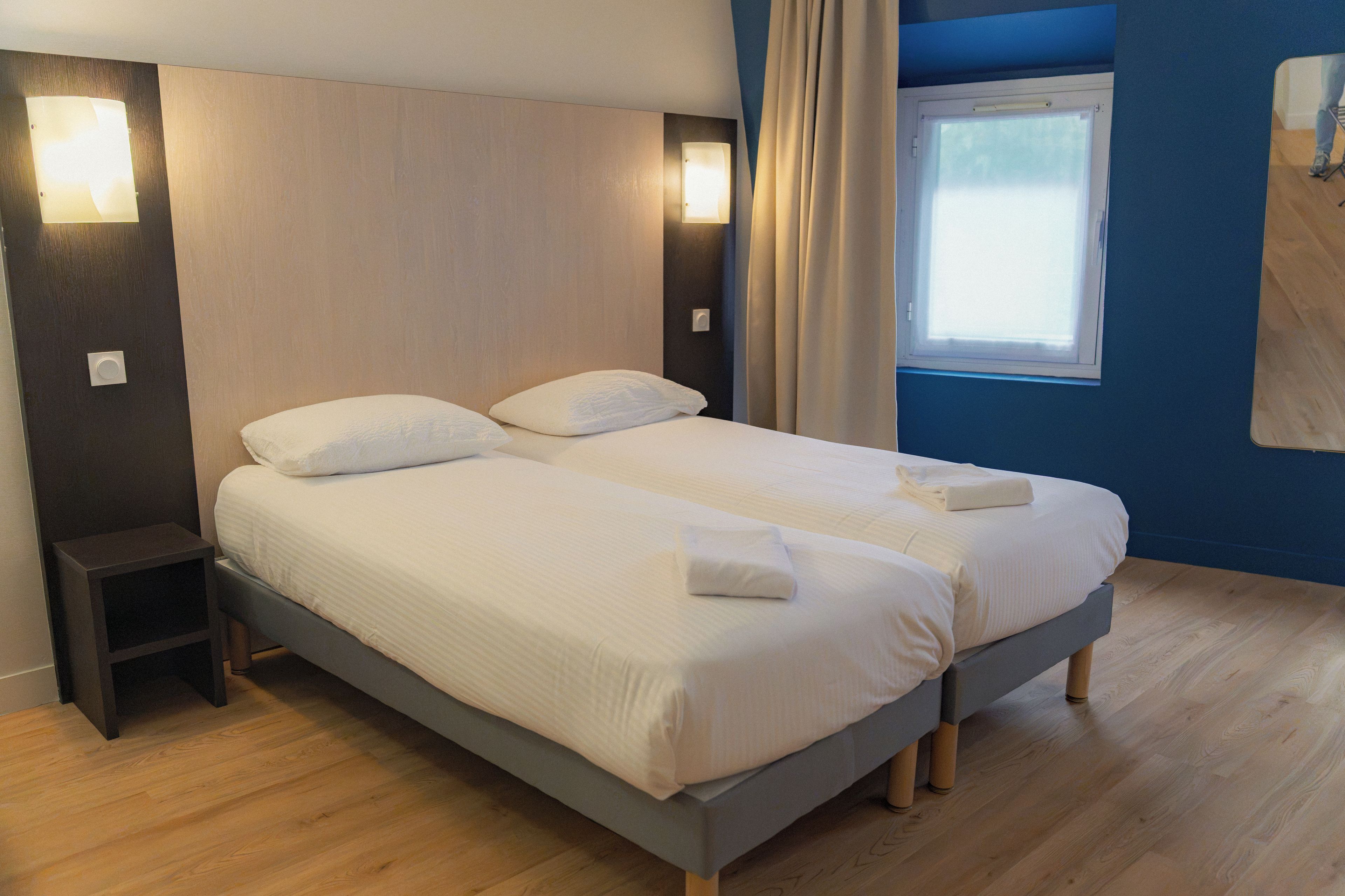 Un lit double avec du linge de lit blanc et des oreillers, une table de chevet, une fenêtre avec des rideaux et un luminaire fixé au mur.