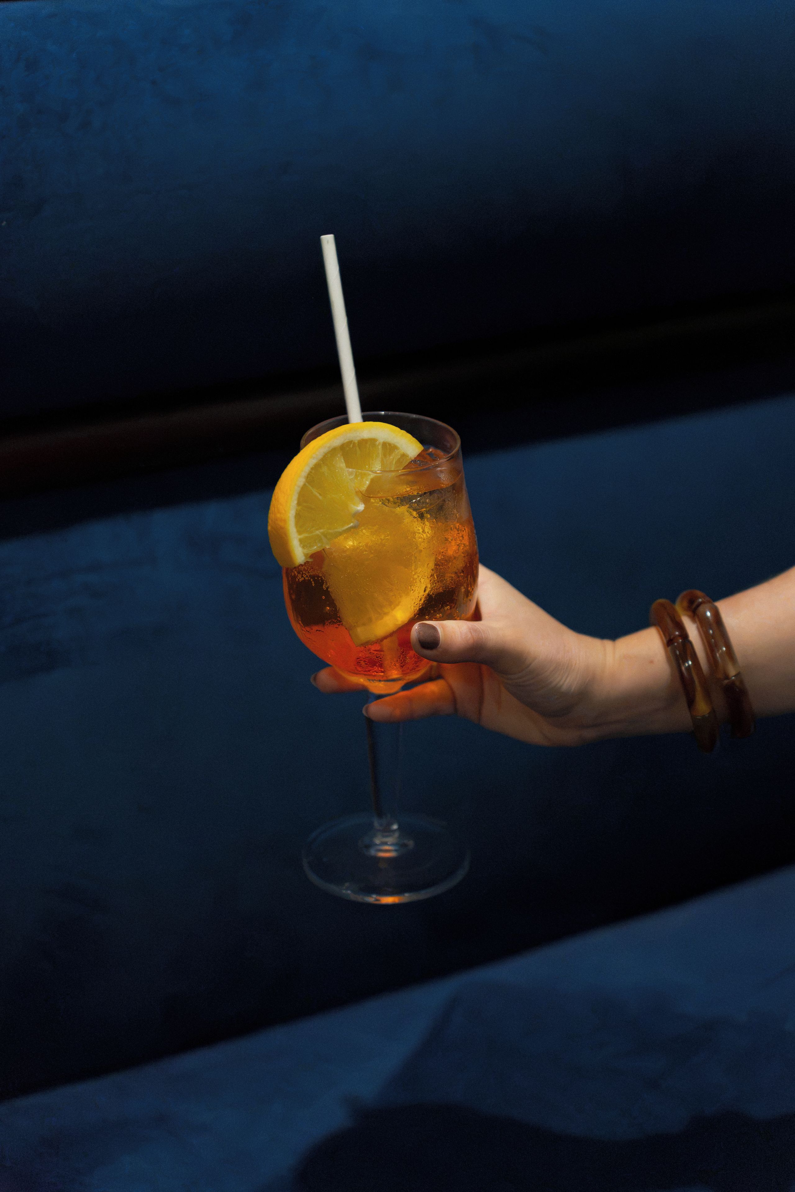 A hand holding a cocktail glass with a straw, containing a beverage and an orange slice garnish.