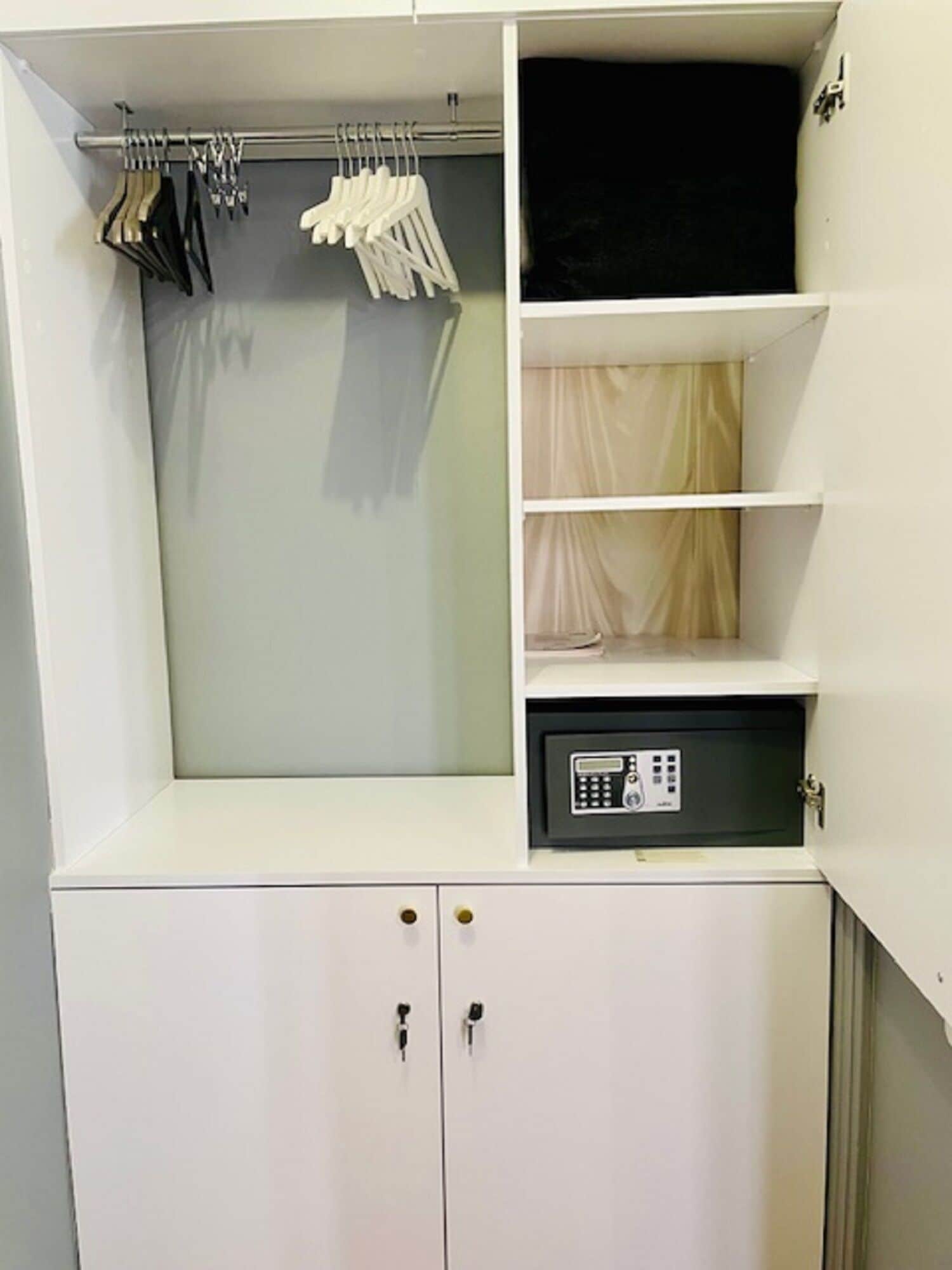 Ein weißer Kleiderschrank mit zwei Türen, eine Kleiderstange mit Kleidung und ein schwarzer Safe.