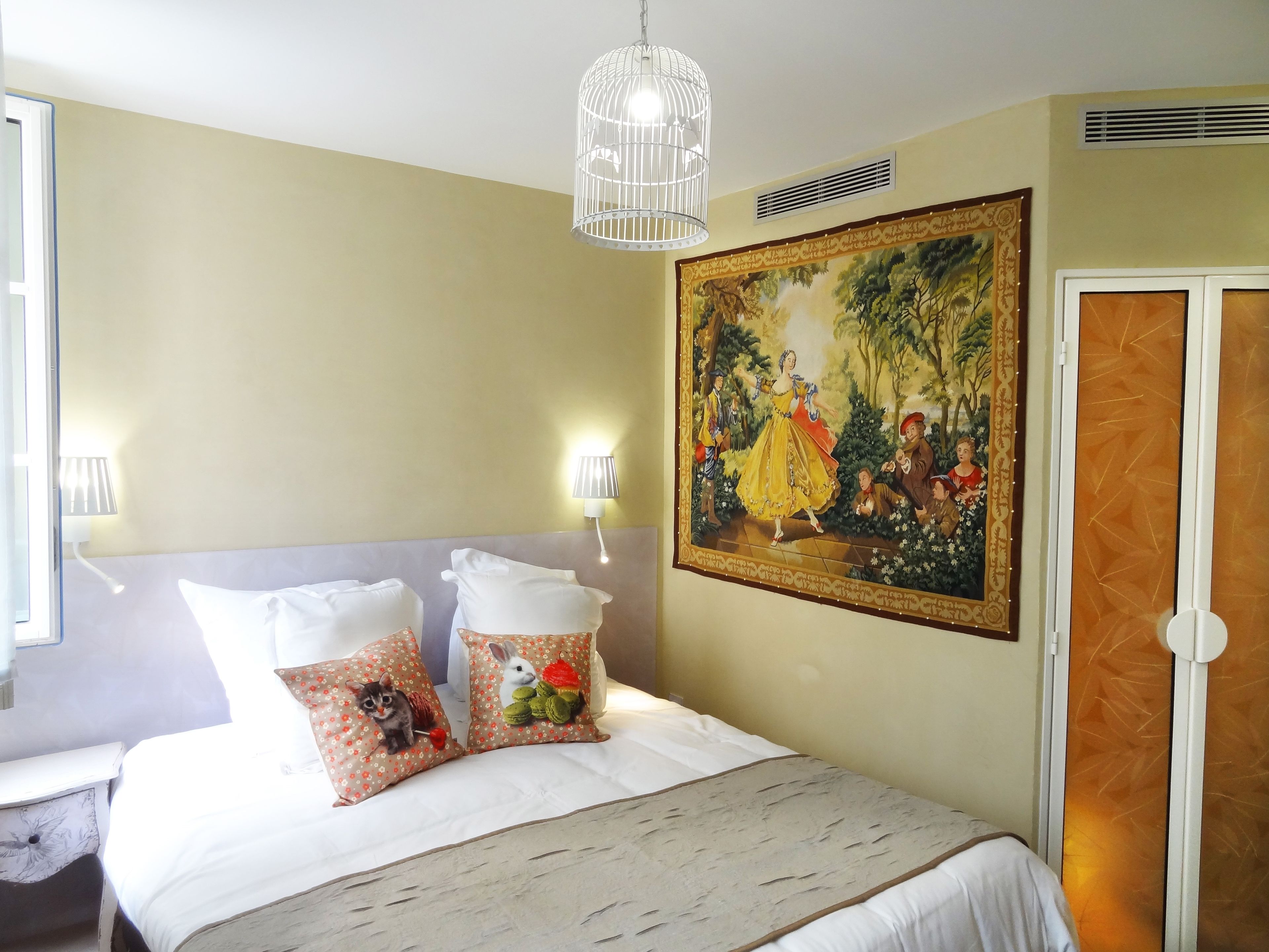 Une chambre d’hôtel avec un lit, deux oreillers ornés de motifs représentant des lapins, un tableau représentant une femme en robe jaune, un luminaire en forme de cage à oiseaux blanche et une porte dotée d’un panneau décoratif.