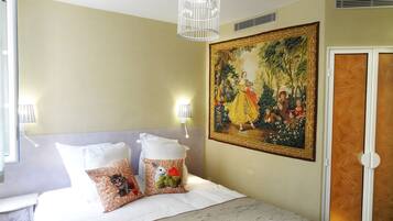 Une chambre d’hôtel avec un lit, deux oreillers ornés de motifs représentant des lapins, un tableau représentant une femme en robe jaune, un luminaire en forme de cage à oiseaux blanche et une porte dotée d’un panneau décoratif.