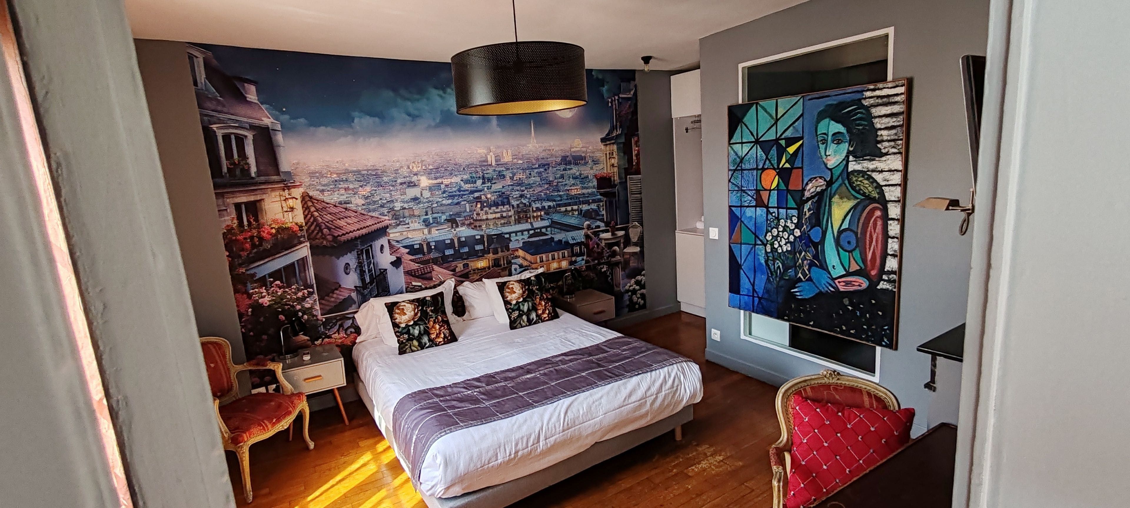 Quarto com mural de paisagem urbana, cama com cabeceira acolchoada, poltrona vermelha estofada e um quadro na parede.