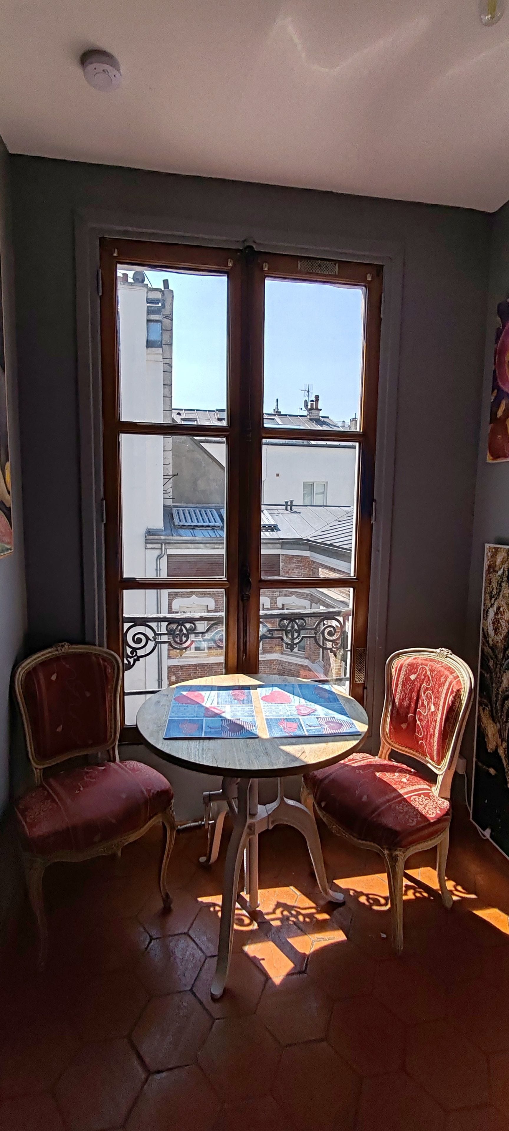 A room with a round table, two chairs, and a view of buildings through large windows.