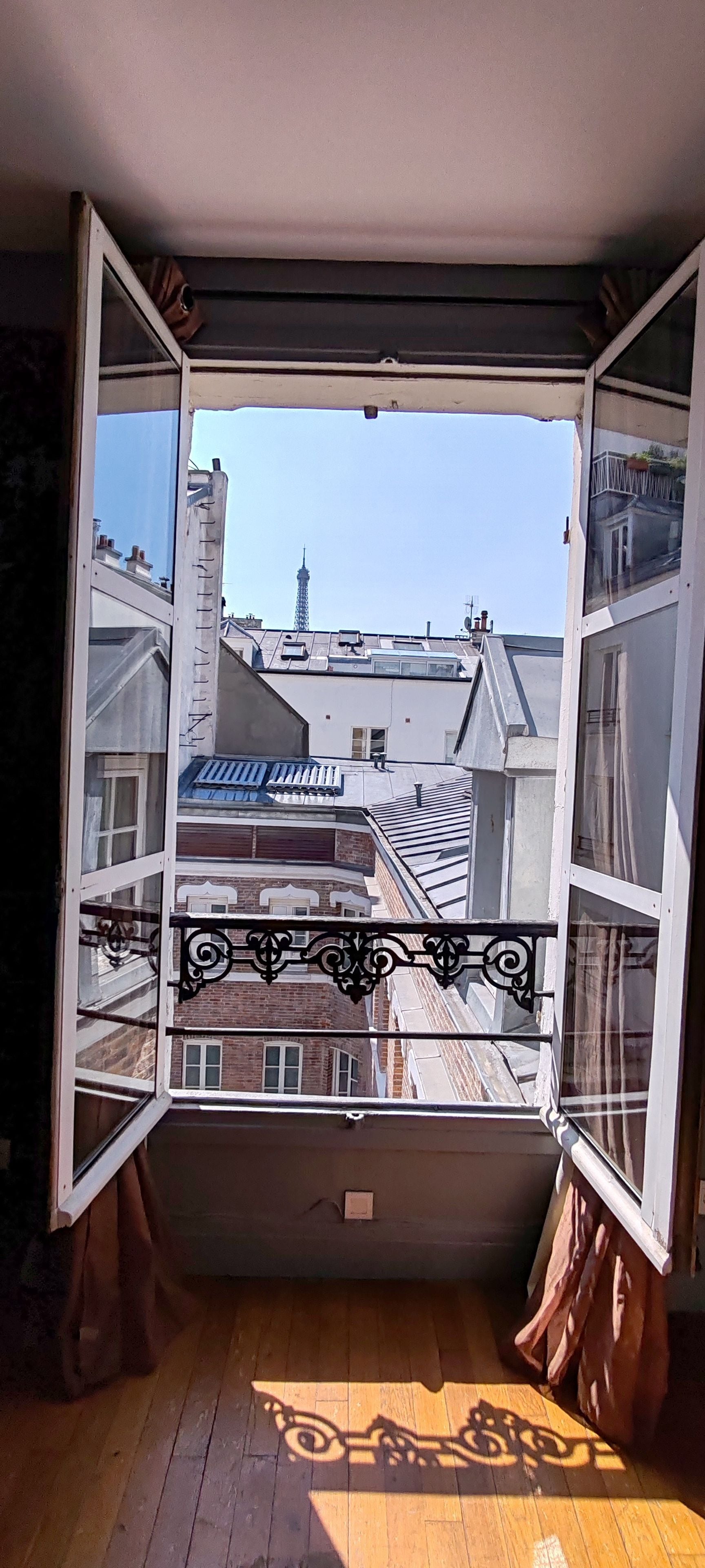 A room with a large open window, wooden flooring, and a view of neighboring buildings and a tower.