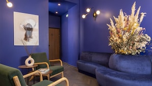 Een kamer met een blauwe wand, een houten deur, een blauwe bank, een groene fauteuil, een kleine tafel met een vaas, en een wandlamp.