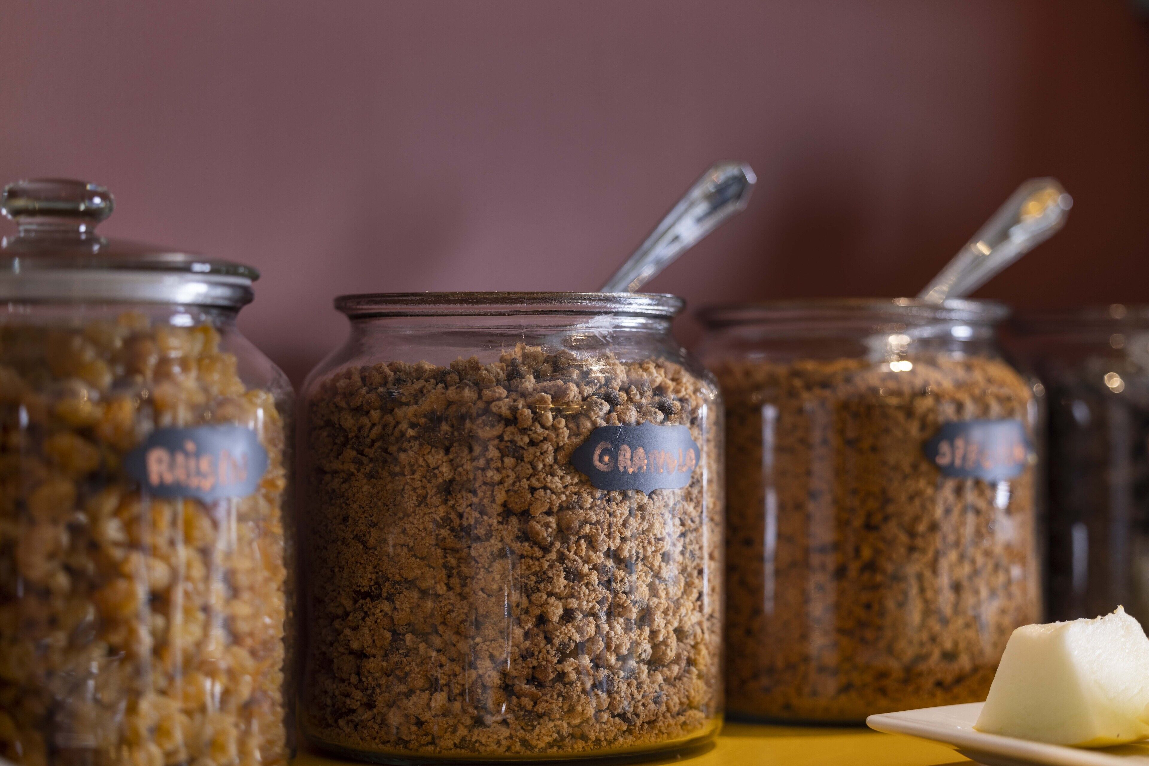 Three glass jars labeled "Granola" with a spoon in one, and a piece of butter on a plate.