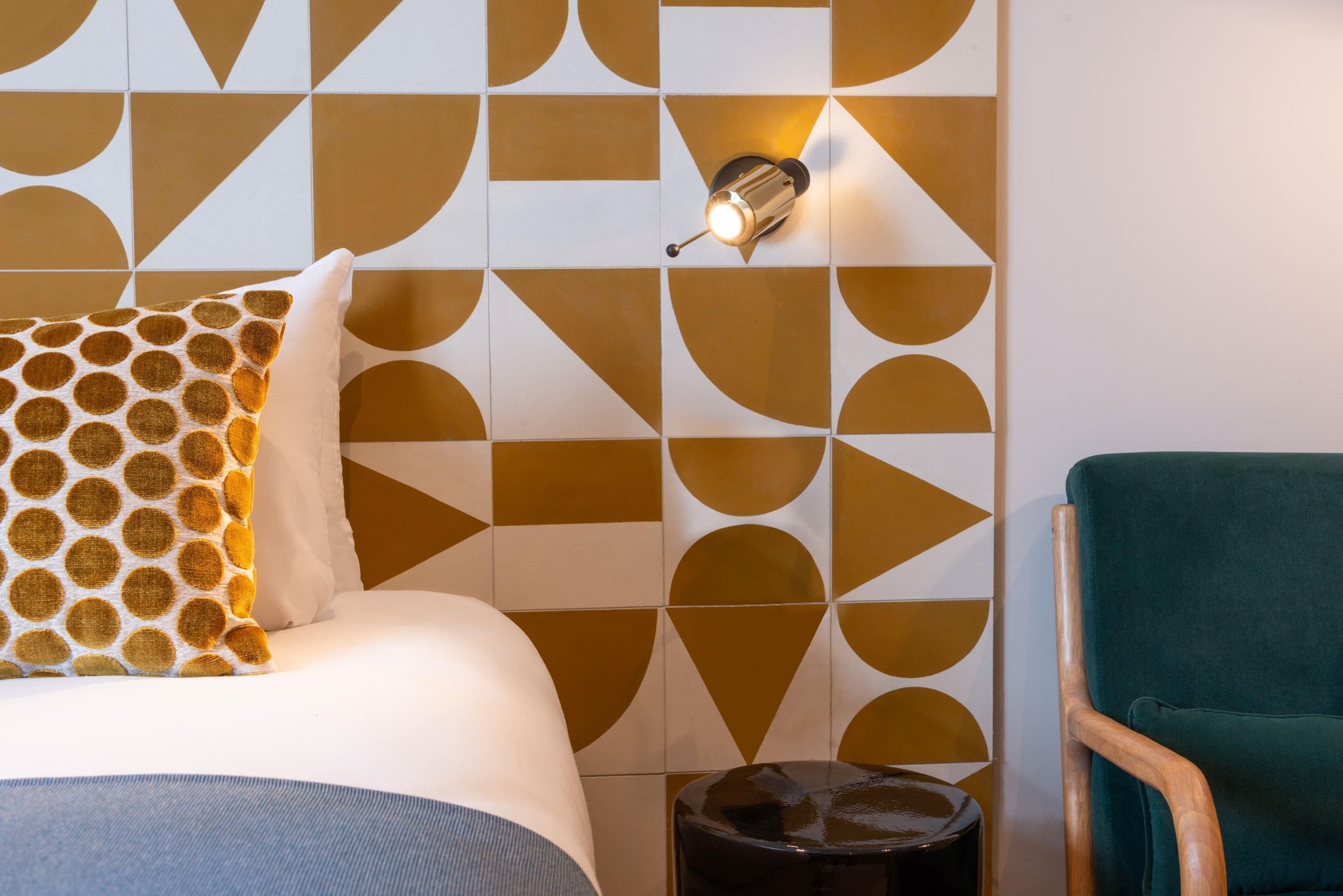 Een netjes opgemaakt bed met een gekleurd kussen, een muur met een geometrisch tegelpatroon, een wandlamp en een groene fauteuil.
