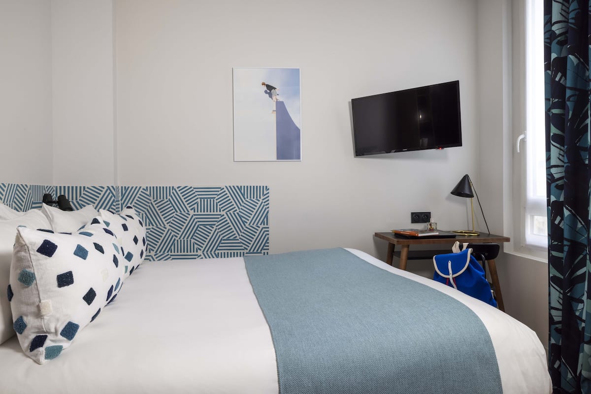 Ein ordentlich eingerichtetes Hotelzimmer mit einem Bett, einem wandmontierten Flachbildfernseher, einem Schreibtisch mit Lampe und einem fenster mit Vorhängen.