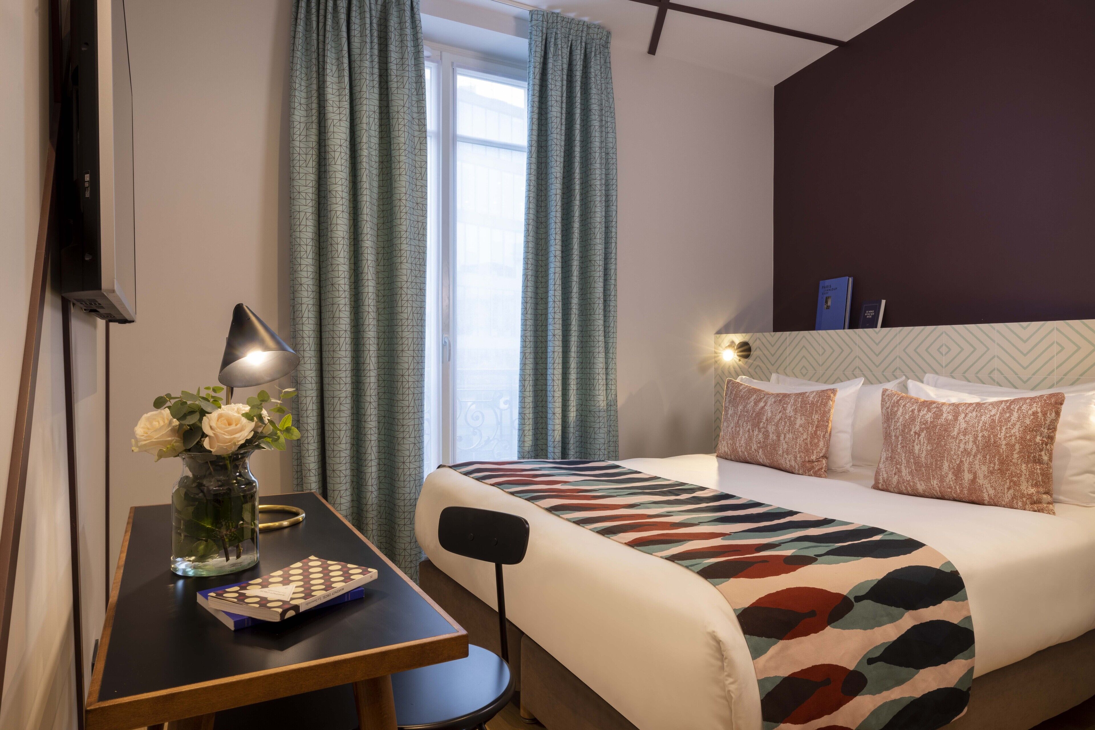 A hotel room with a bed, a desk with a lamp, a vase with flowers, and a window with curtains.