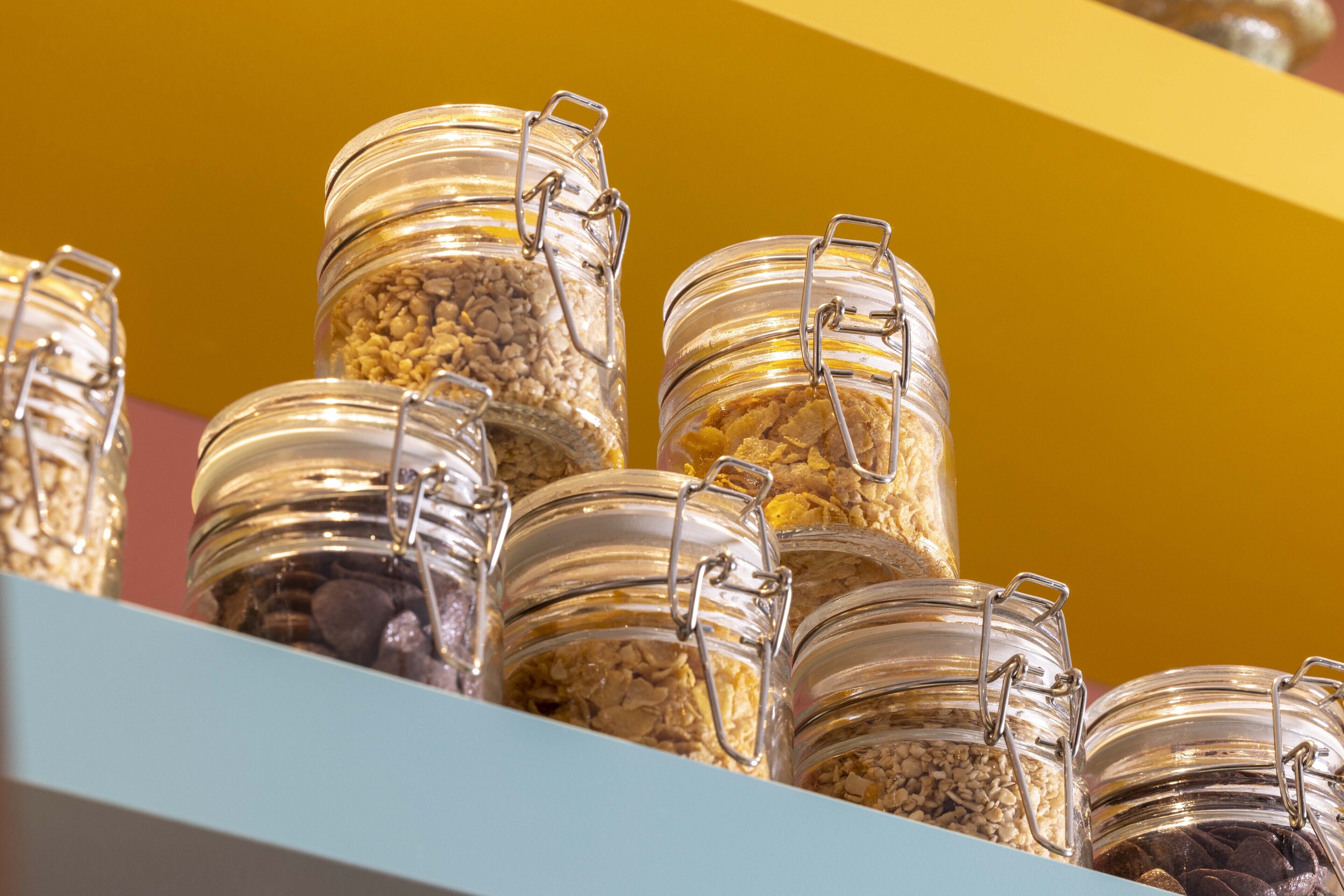A collection of glass jars with metal clasps, containing various grains and seeds, displayed on a shelf against a yellow background.