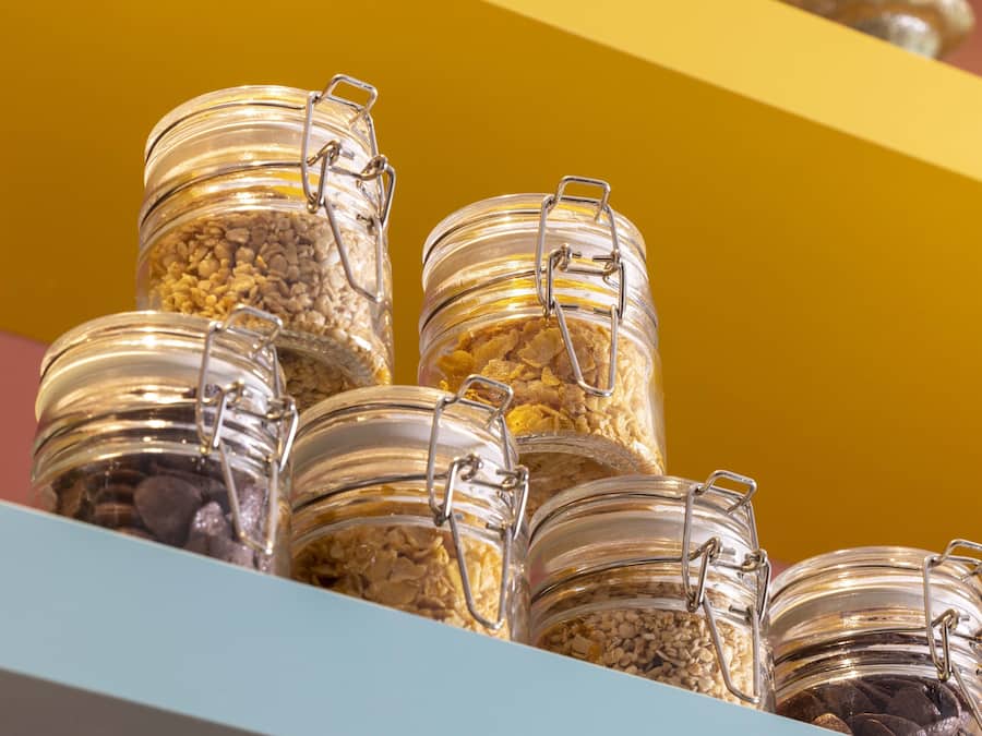 A collection of glass jars with metal clasps, containing various grains and seeds, displayed on a shelf against a yellow background.