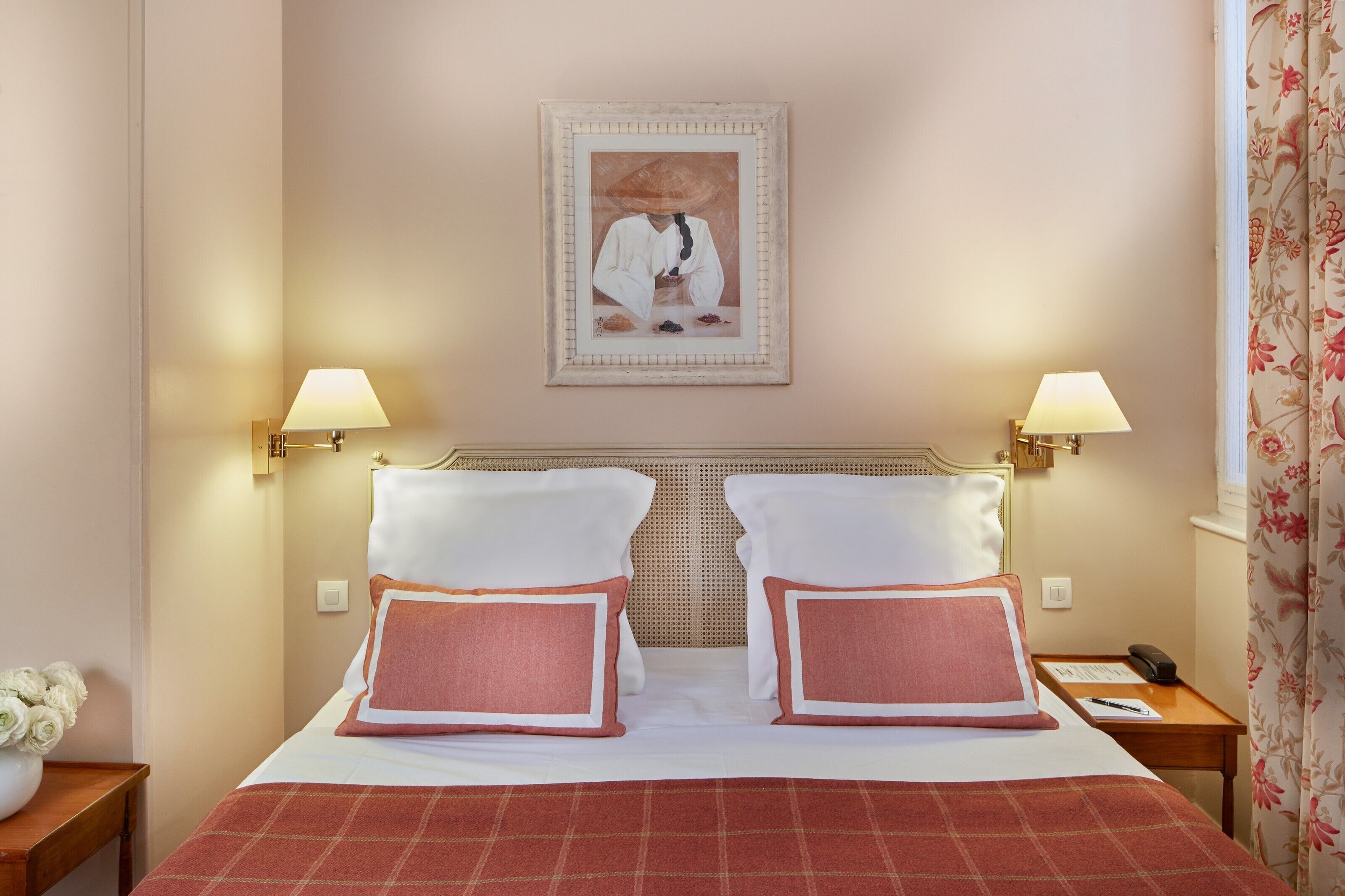 A hotel room with a bed, two bedside tables, a nightstand, a framed picture on the wall, and a floral curtain.
