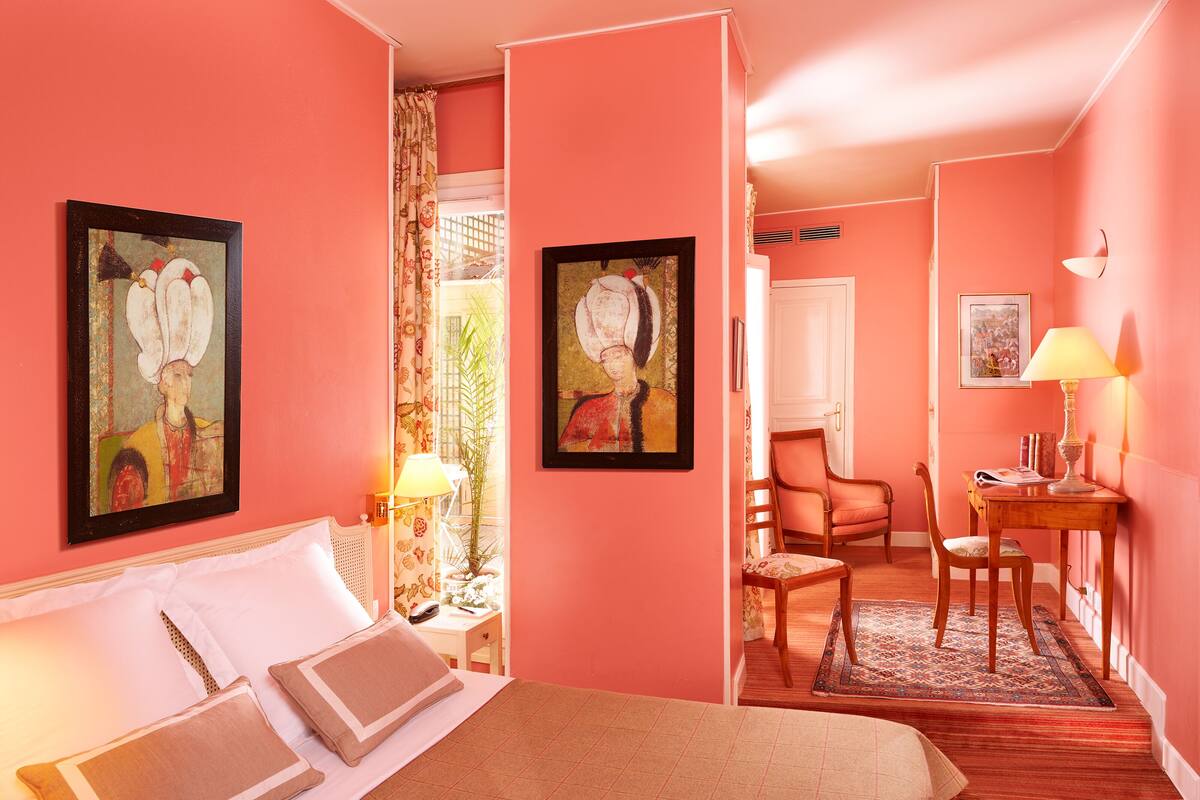 A pink-themed hotel room with a bed, a desk, a chair, and two framed artworks on the walls.