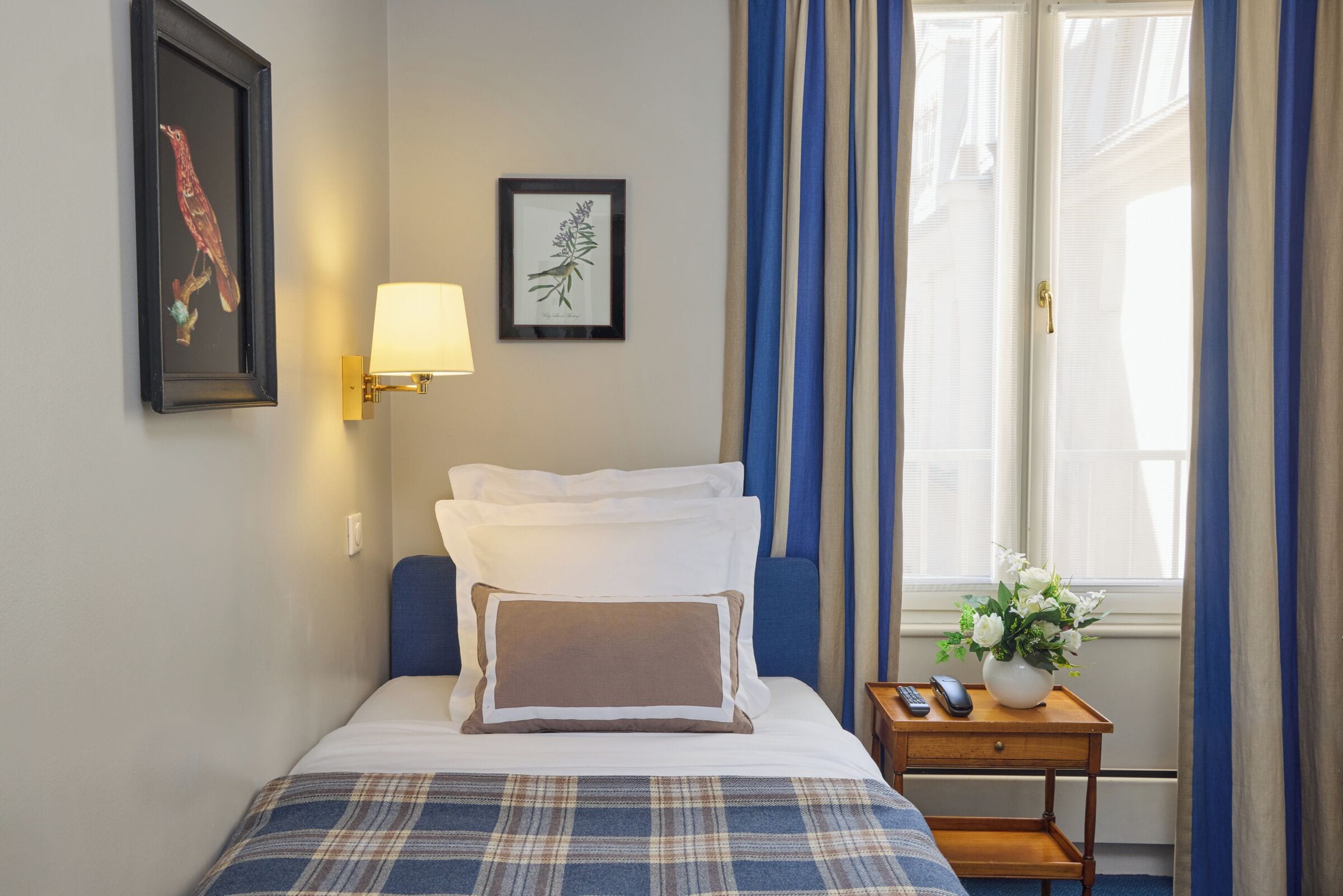 A hotel room with a bed, a bedside table with a lamp, a framed bird and plant on the wall, and a window with curtains.