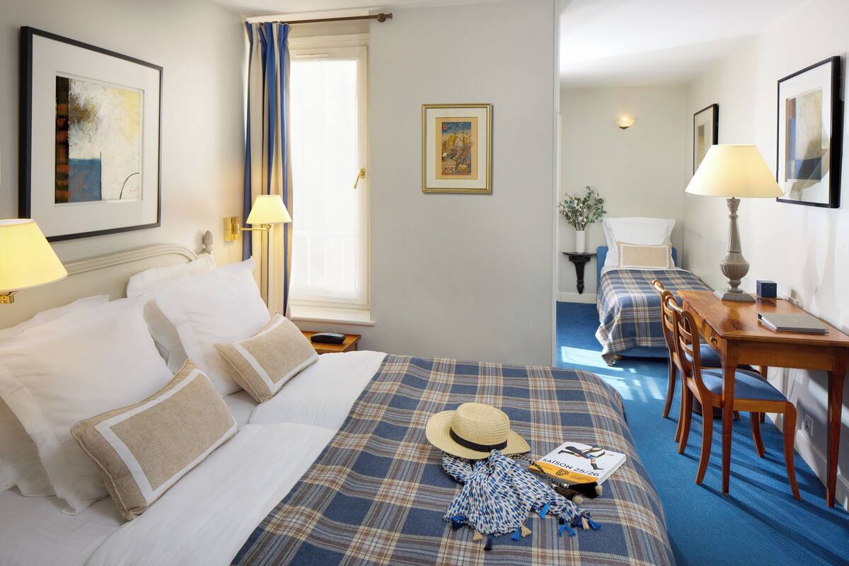 A hotel room with a bed, a desk, and a chair. There are two lamps on the desk and a window with curtains.