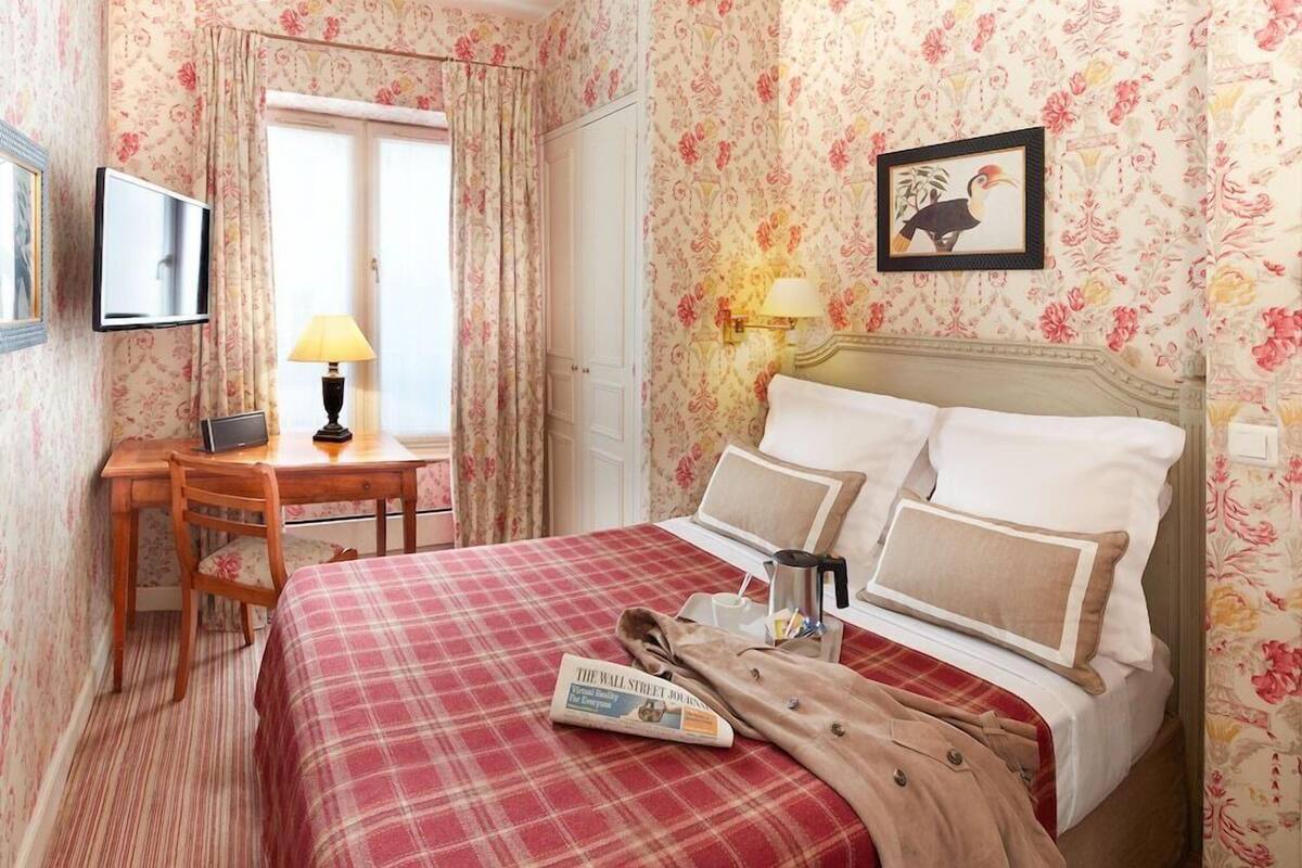 A hotel room with floral wallpaper, a bed with a red plaid bedspread, a desk with a lamp, and a framed picture on the wall.