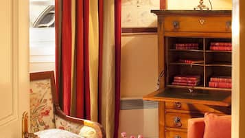 Een gezellige kamer met een bloemen fauteuil, een rode bank, een houten kast vol boeken, een tafel met een bloevas en een raam met gordijnen.