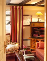 A cozy room with a floral armchair, a red sofa, a wooden cabinet with books, a table with a vase of flowers, and a window with curtains.