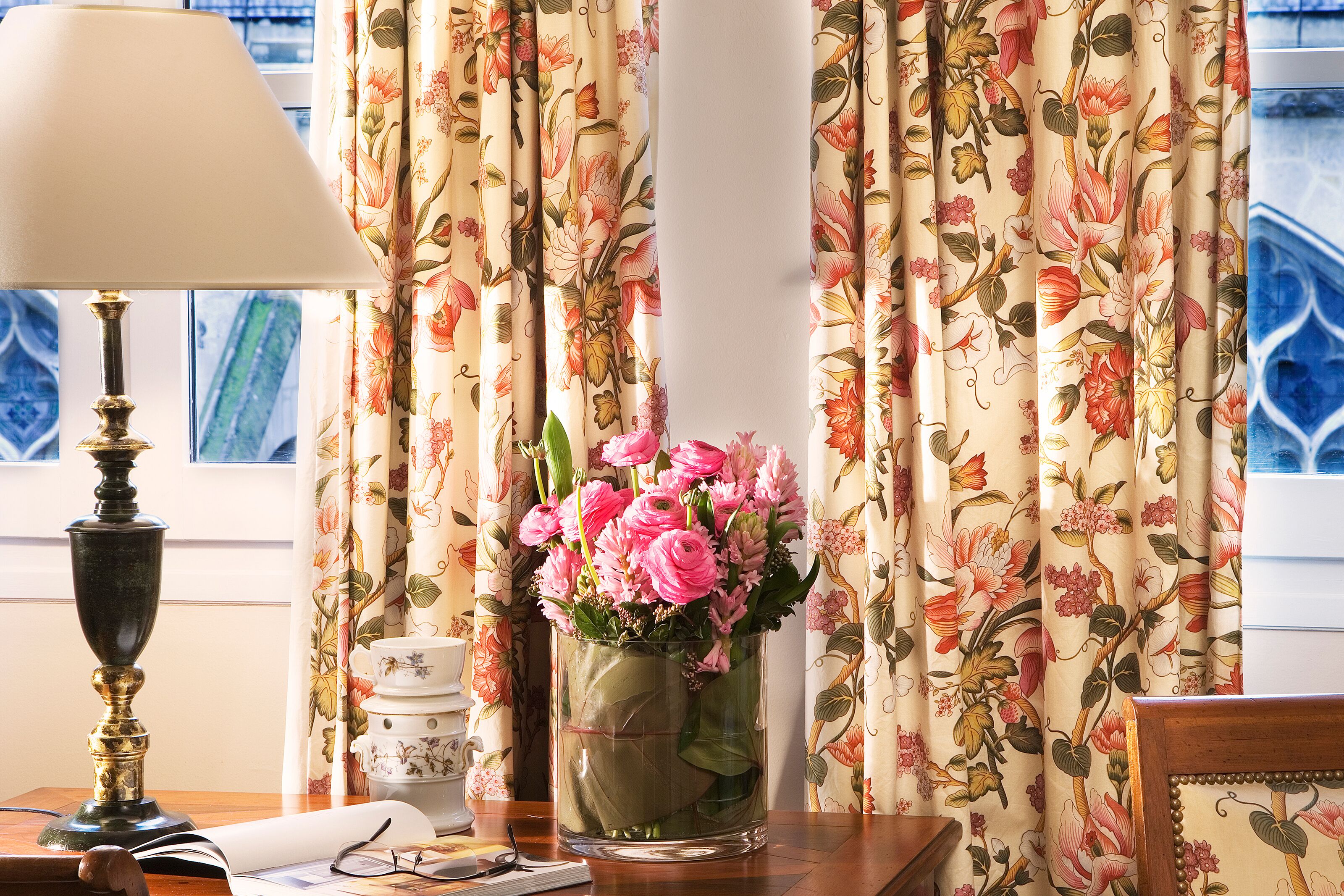 A room with a floral curtain, a vase with pink flowers, a lamp, and a window with a view.