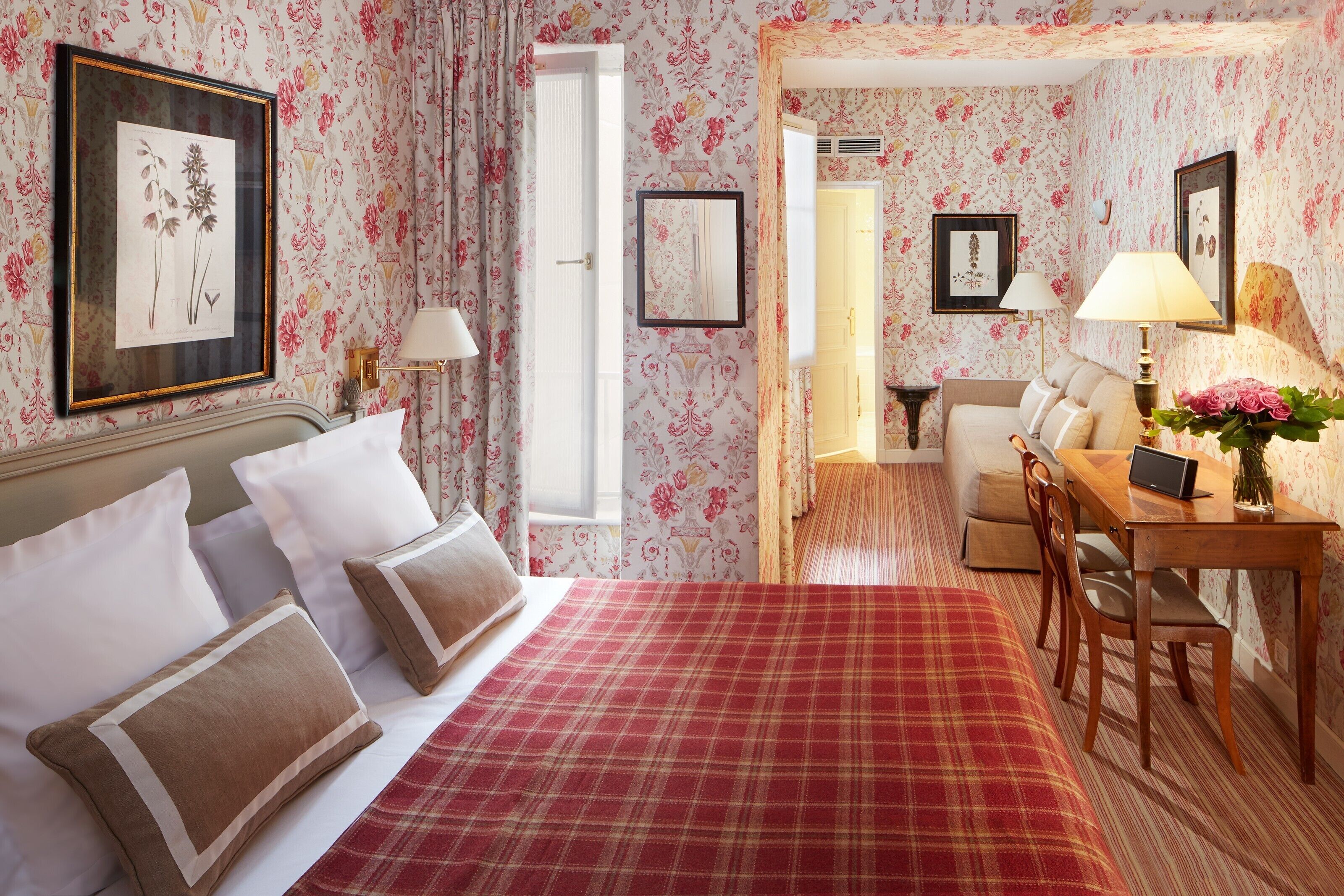 A hotel room with a bed, a desk, and a chair. There is a window and a door leading to another room.