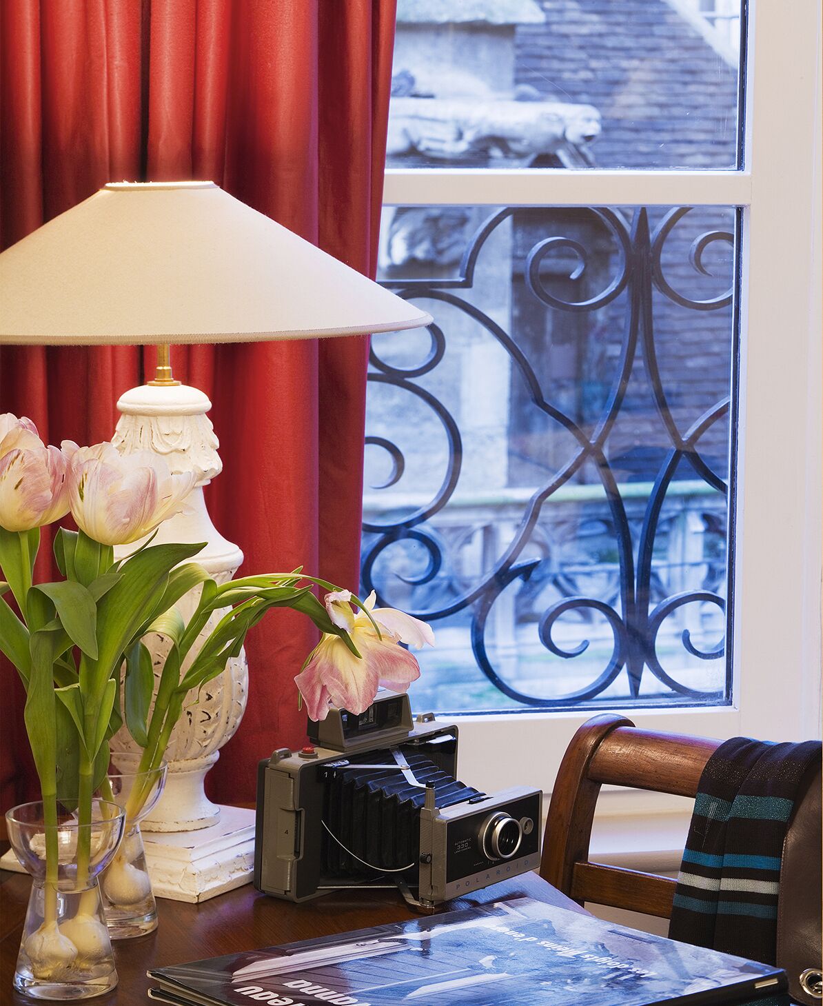 A room with a table featuring a vintage Polaroid camera, a lamp, and a vase with flowers.