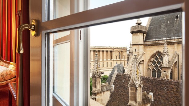A view from a window with a wooden frame and a brass handle, overlooking a historic building with Gothic architecture and a brick wall. - Relais Du Louvre (Paris)