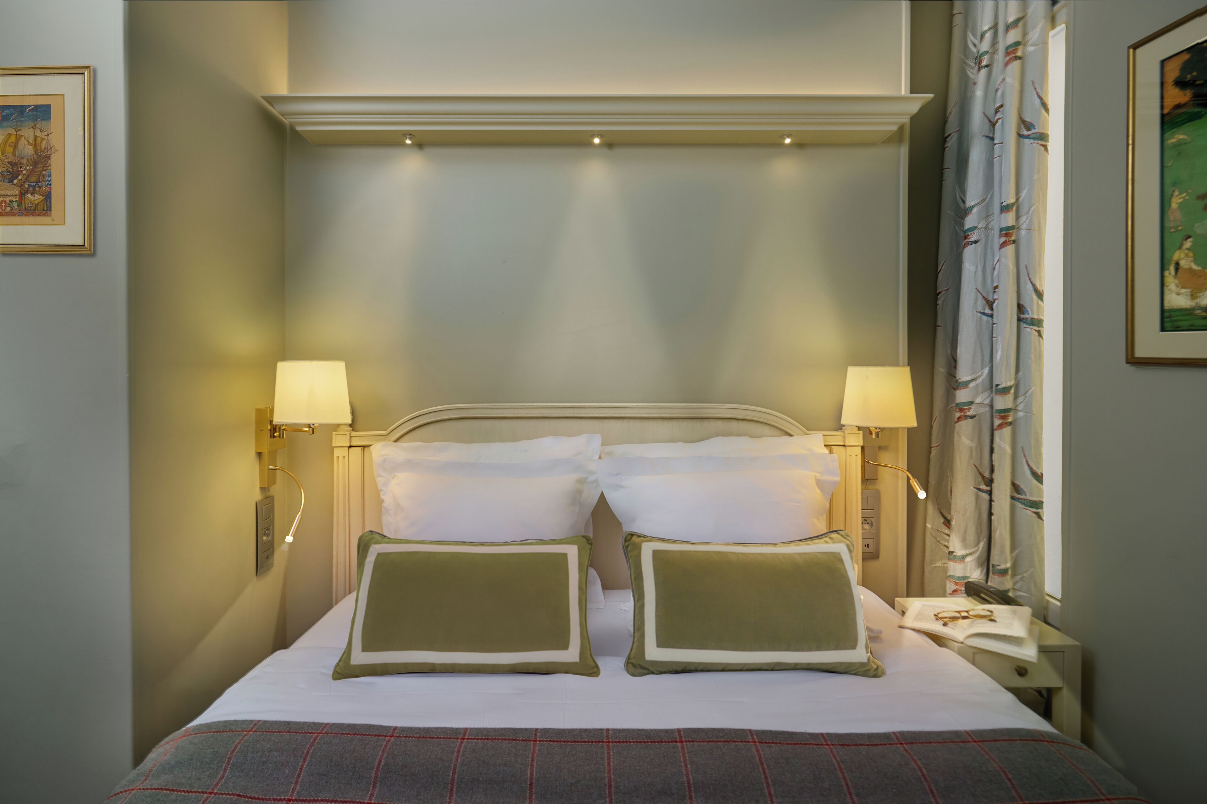 A neatly made bed with two pillows, two bedside lamps, and a nightstand with a book and glasses.