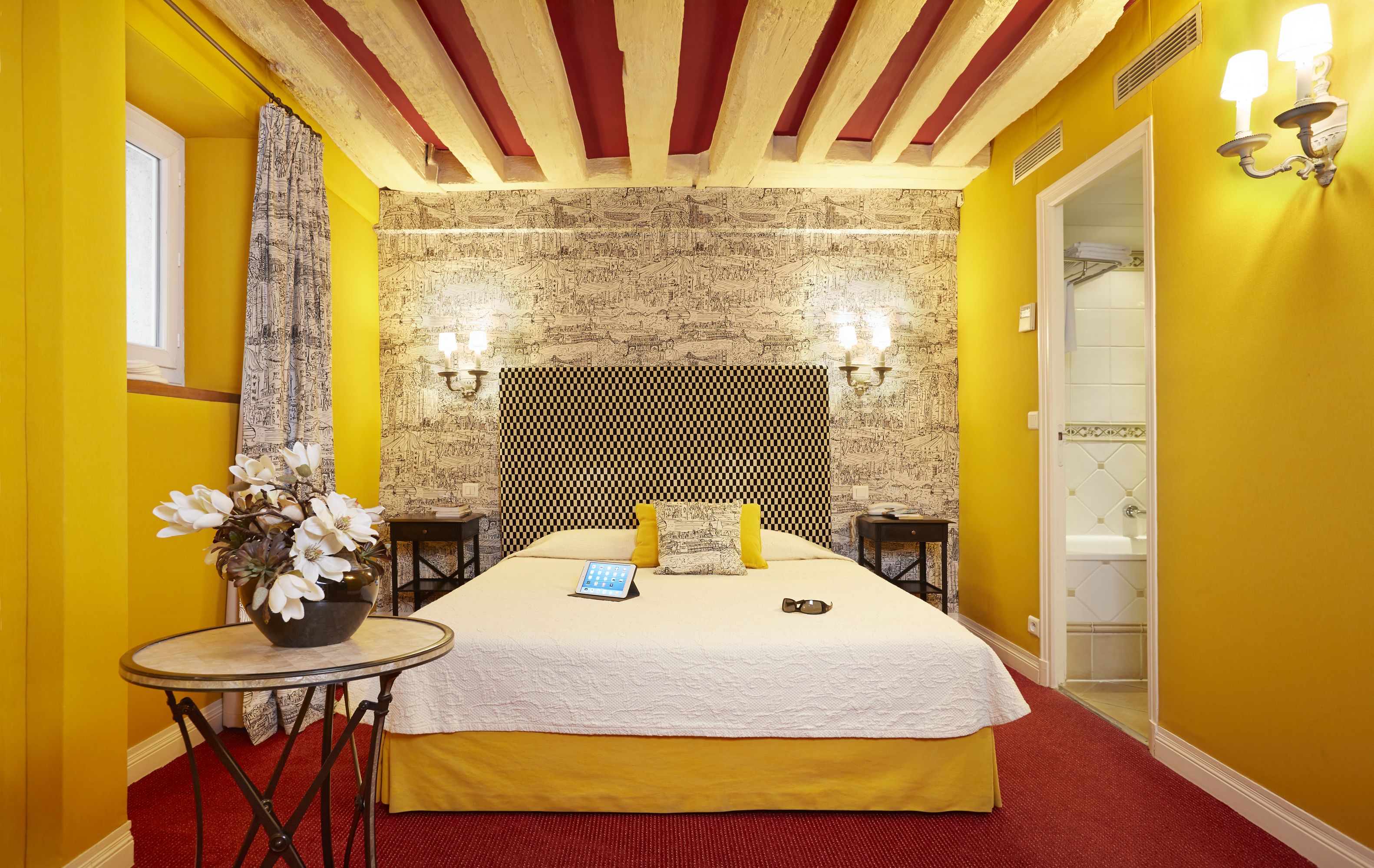 Una habitación de hotel con una cama, una mesita con un florero y una alfombra roja, además se ve un baño a través de una puerta abierta.