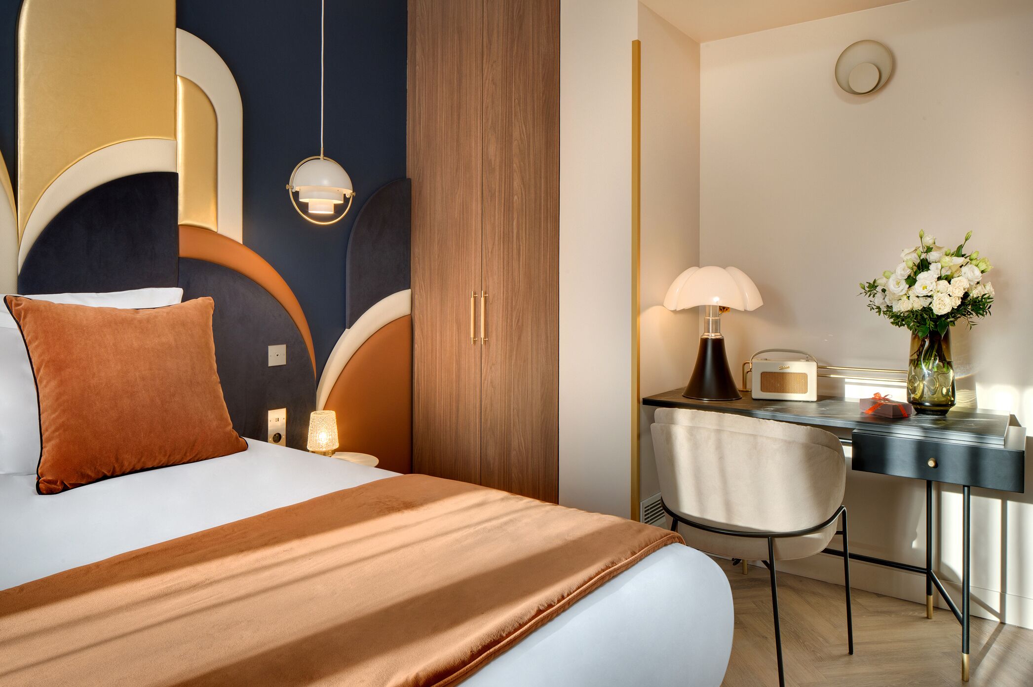 Une chambre d’hôtel avec un lit, un bureau, une chaise, une lampe et un vase avec des fleurs.
