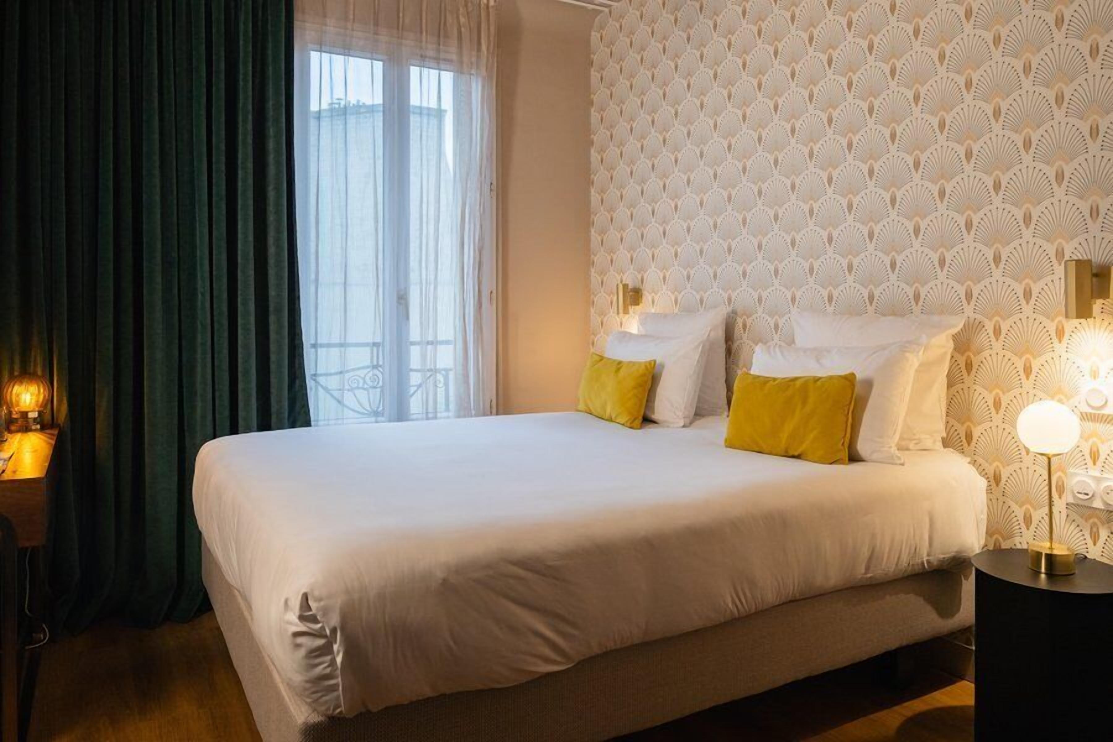 A hotel room with a large bed, a window with curtains, a patterned wall, and a bedside lamp.