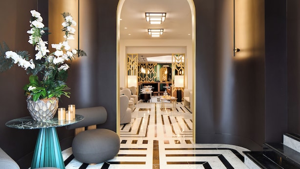 Una hall d'albergo moderna con un pavimento a motivi geometrici, un'area salotto centrale e una pianta decorativa.