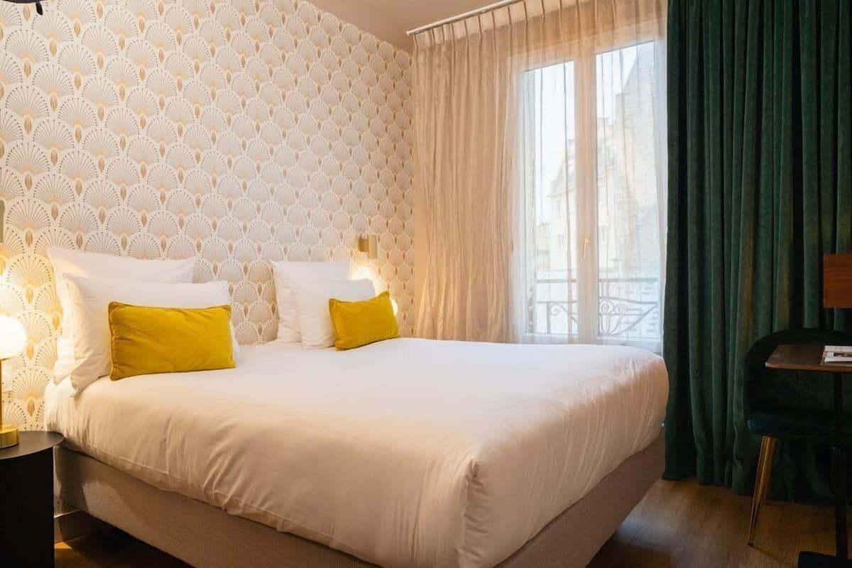 Una camera d'albergo con un letto grande, due lampade sui comodini, una finestra con tende leggere e vista all'esterno.